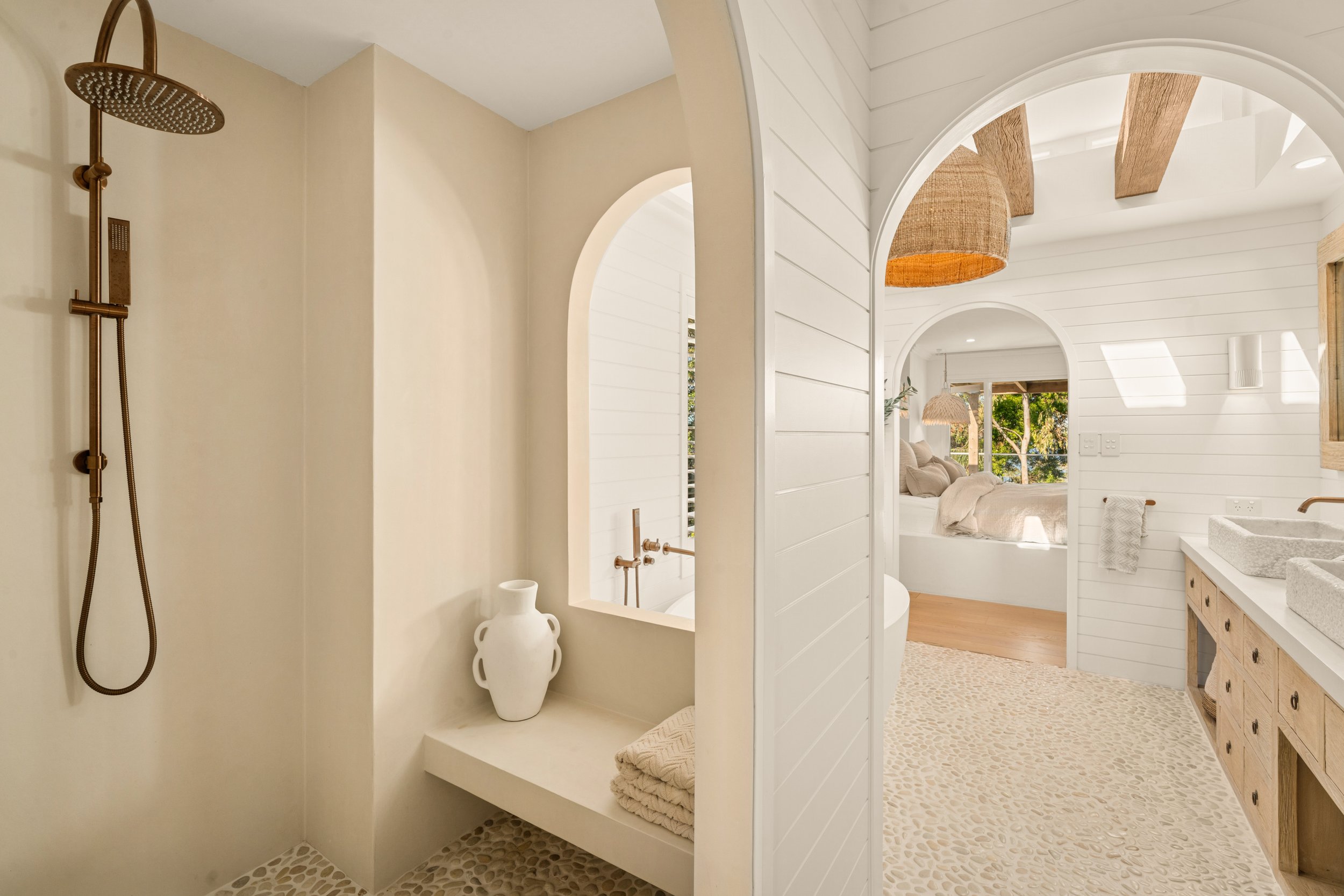 A spacious bathroom with a walk-in shower on the left, a large mirror with a wooden frame, and a vanity with stone sinks on the right. The room has white shiplap walls, natural wood accents, and a view into a bedroom with large windows and greenery o