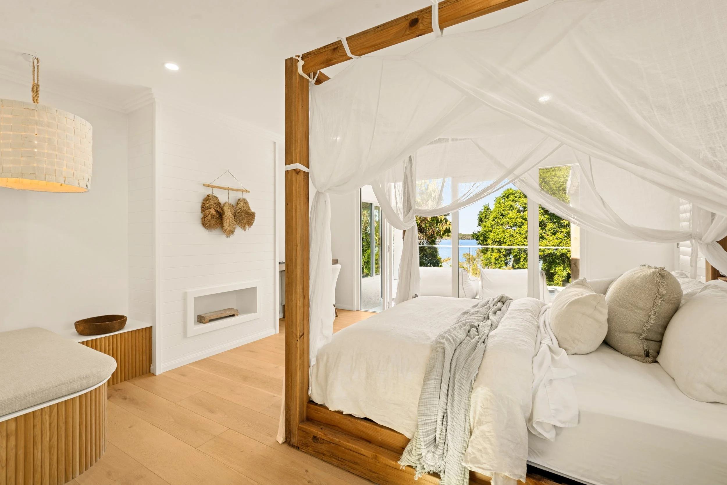 Bright bedroom with white bedding, a canopy bed with draped white curtains, a wooden floor, a balcony with view of greenery and water outside, and minimalistic decor