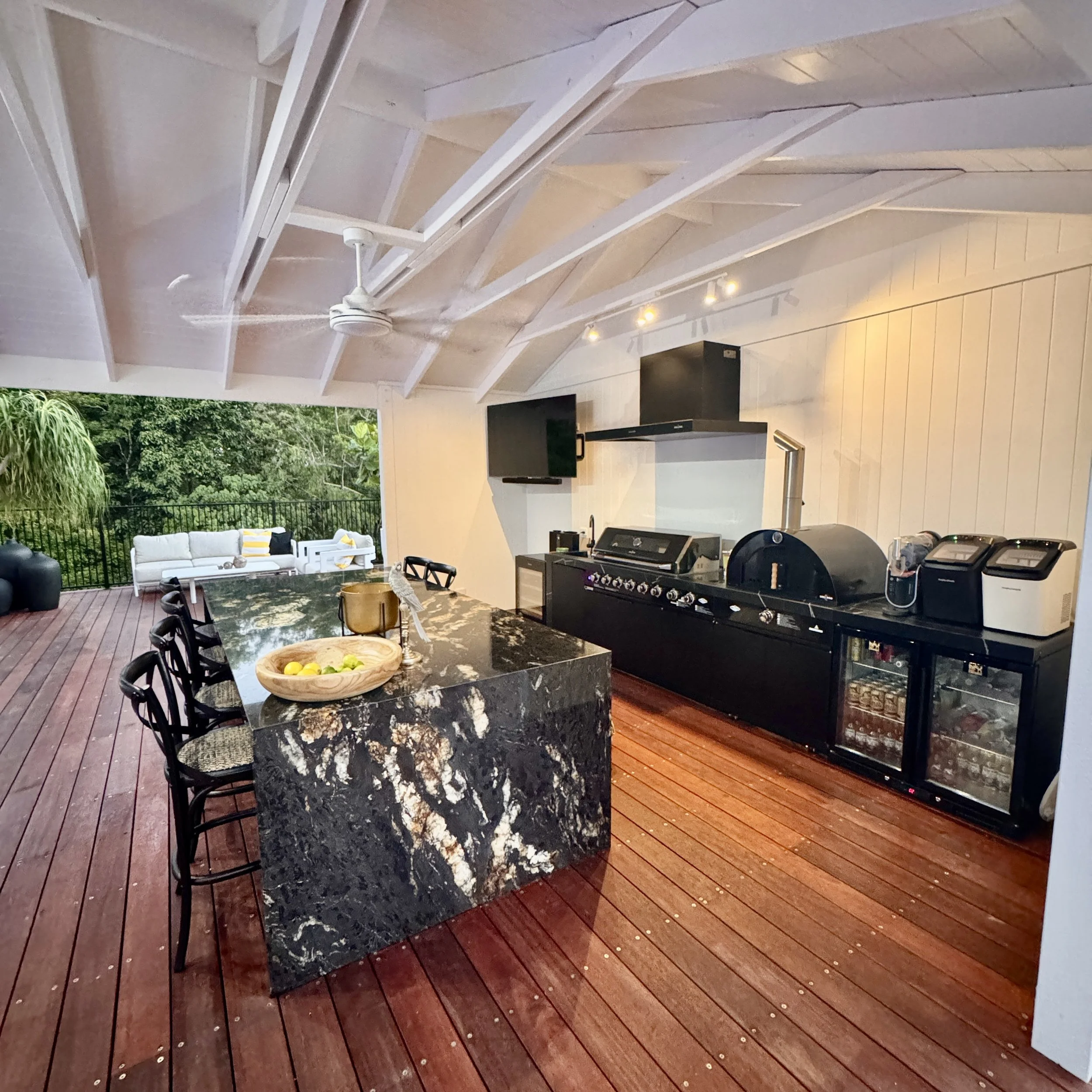 Outdoor patio with wooden decking, black marble kitchen island, grill, and outdoor seating area with white couches and yellow pillows, overlooking lush greenery.