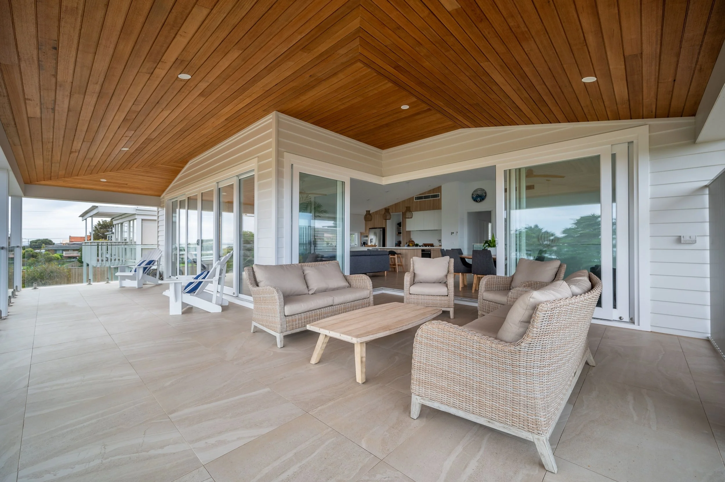 Outdoor patio with wicker lounge chairs and bench, wooden coffee table, overlooking a balcony with white Adirondack chairs, with a view of trees and a neighboring house in the background.