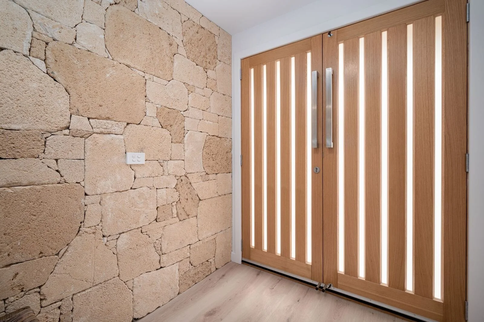A modern wooden door with vertical slats and metallic handles, next to a stone wall with an electrical outlet, in a well-lit room.