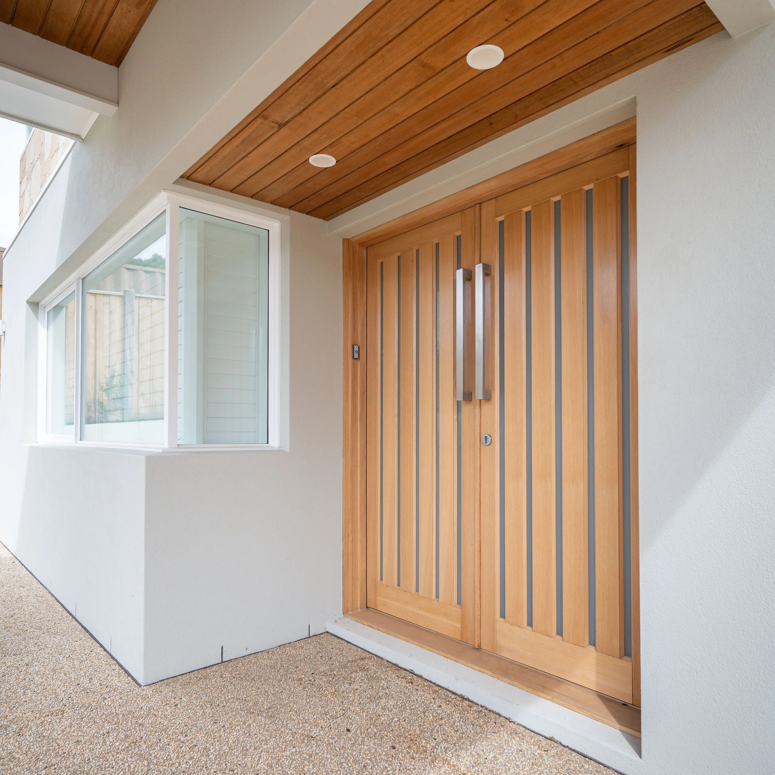 Front view of a modern house entrance with wooden double doors and a large window to the left. The house exterior is painted white, and the porch has a wooden ceiling with recessed lighting.