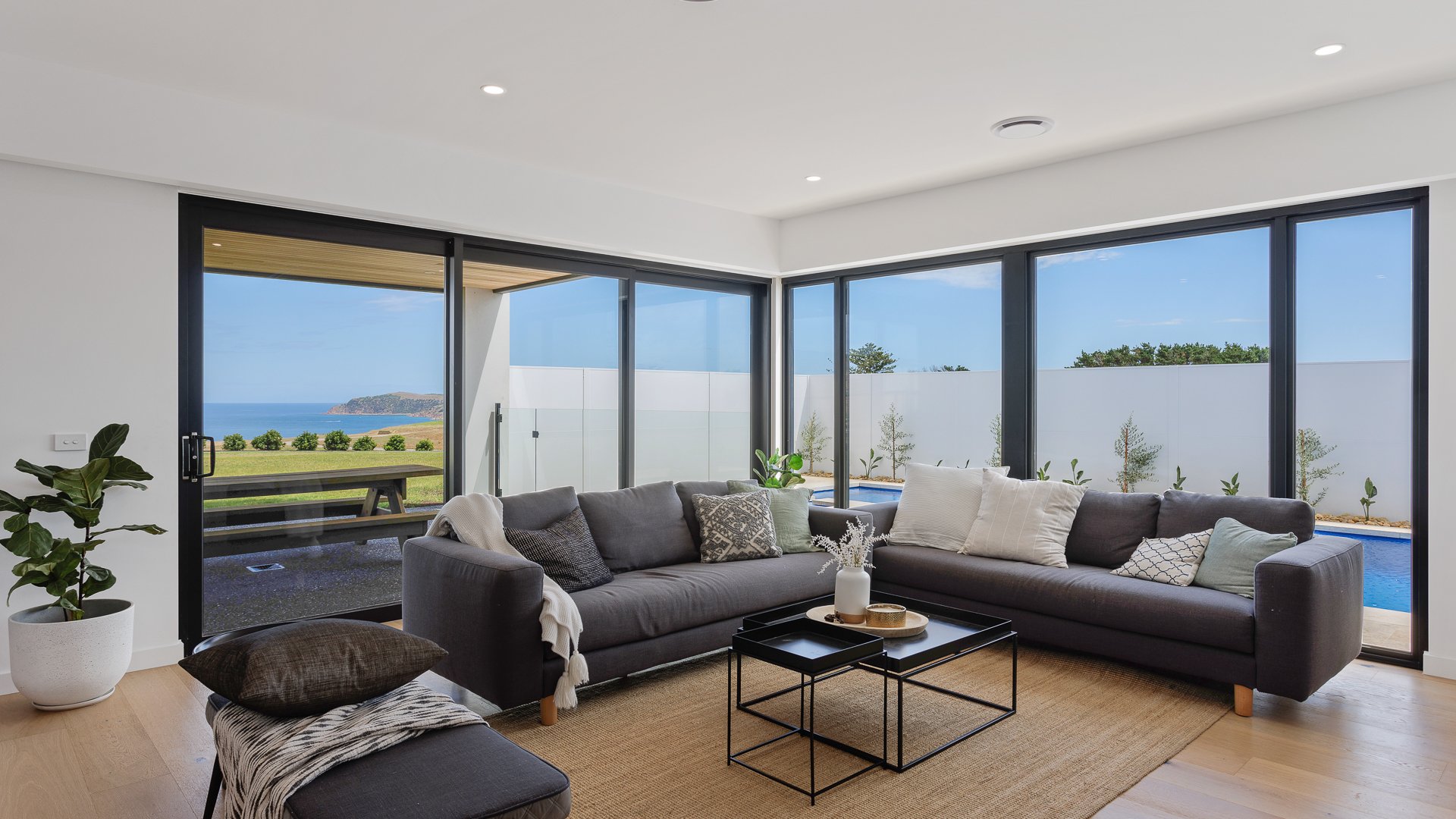 Modern living room with large glass windows showing ocean view, grey sofas with pillows, coffee tables, a potted plant, and a view of the backyard and pool outside.