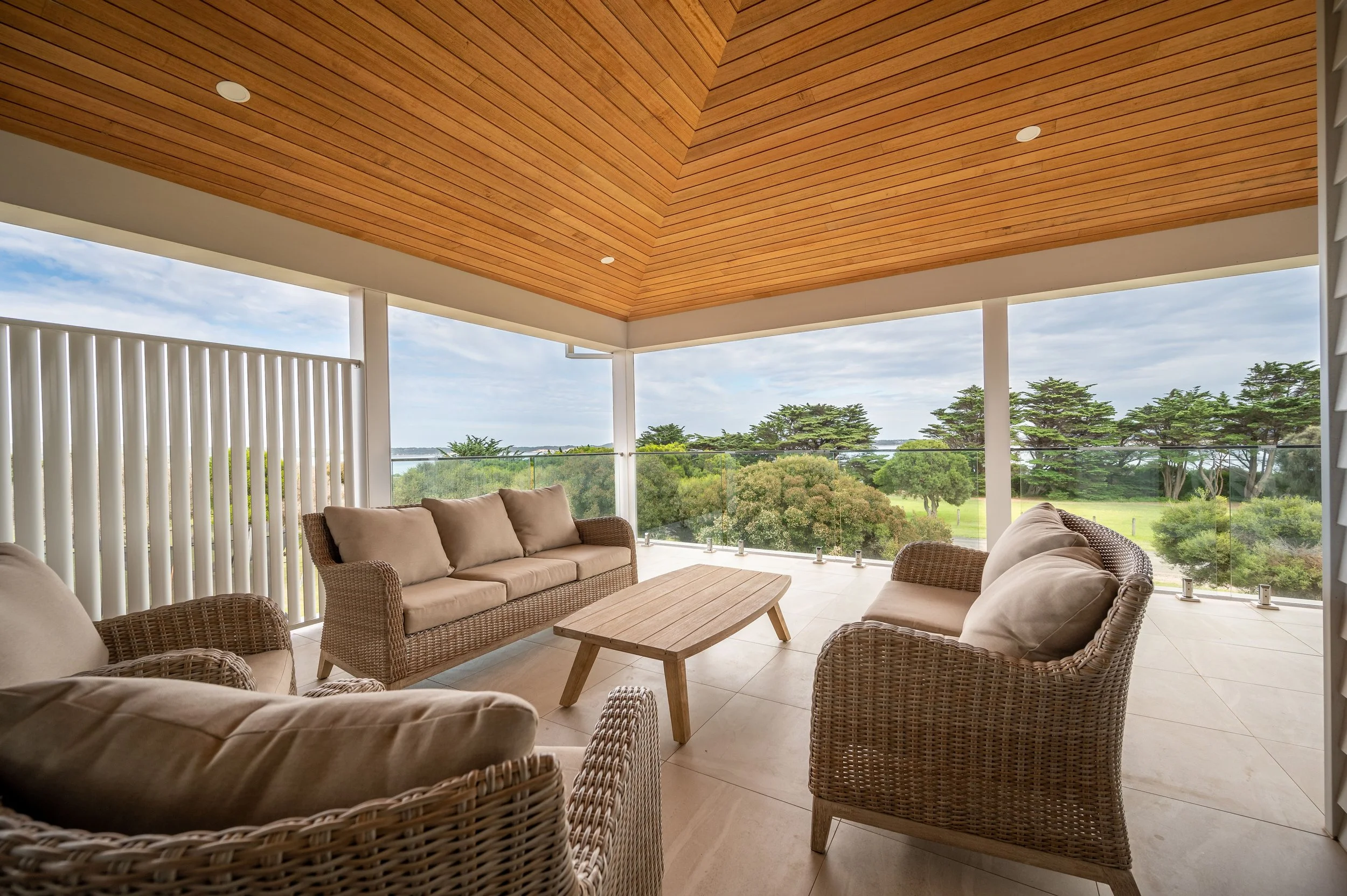 Outdoor covered patio with wicker furniture, beige cushions, and a wooden table surrounded by greenery and trees outside.