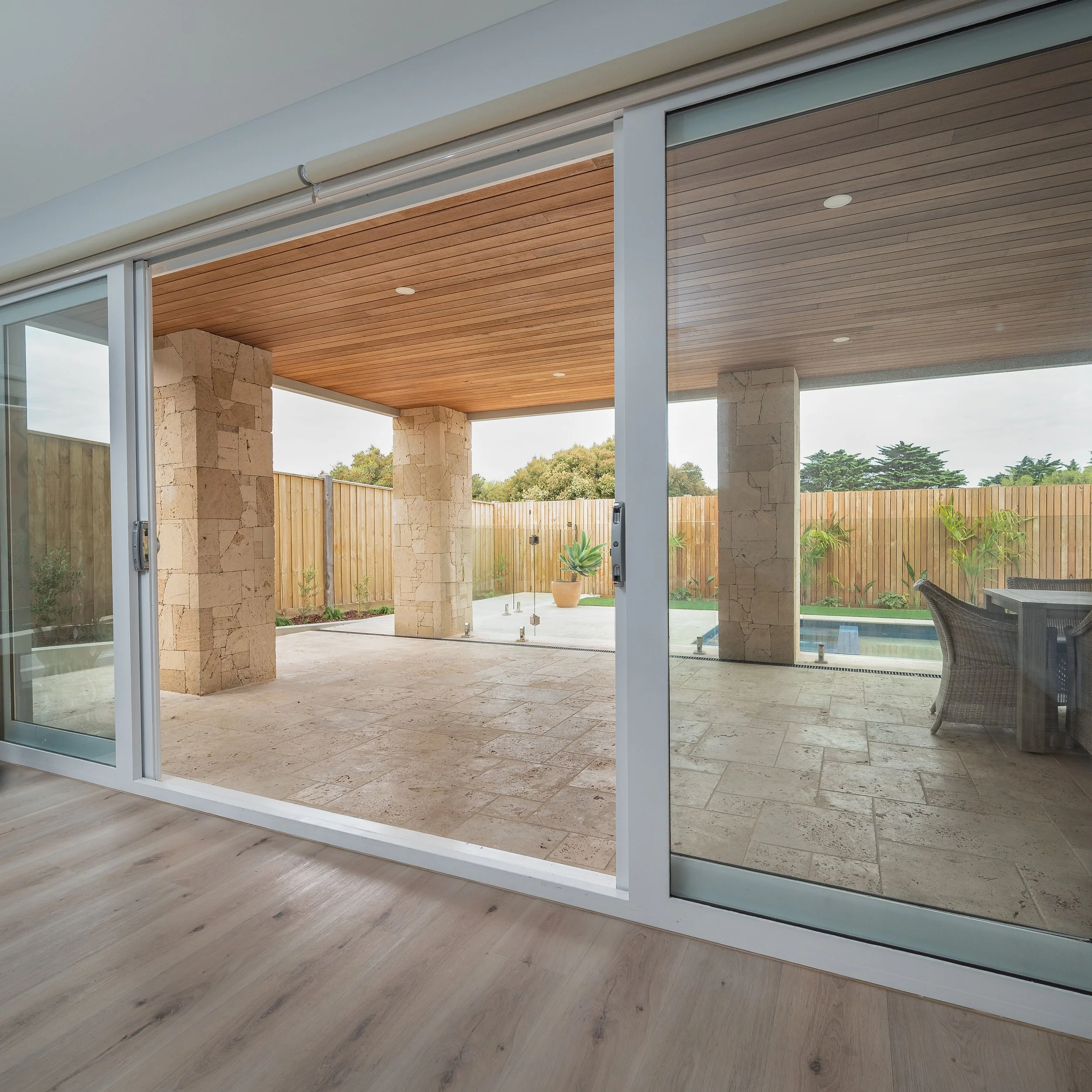 View of a covered patio with stone columns, wooden ceiling, sliding glass doors, and outdoor furniture overlooking a backyard with a pool and garden.