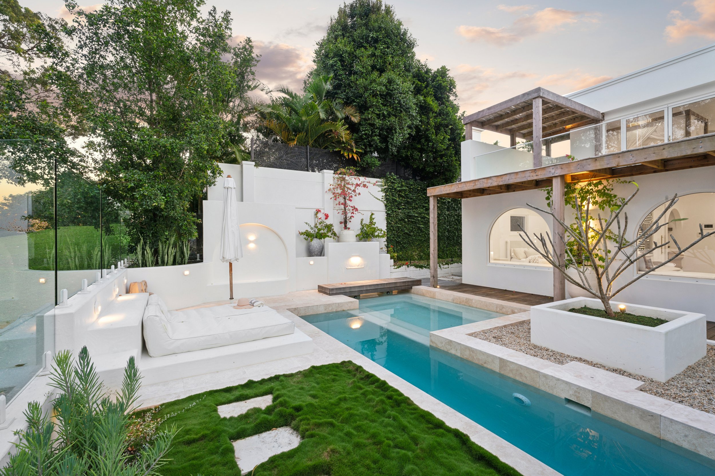 Modern backyard with a small rectangular pool, white walls, greenery, and outdoor lounge area at sunset.