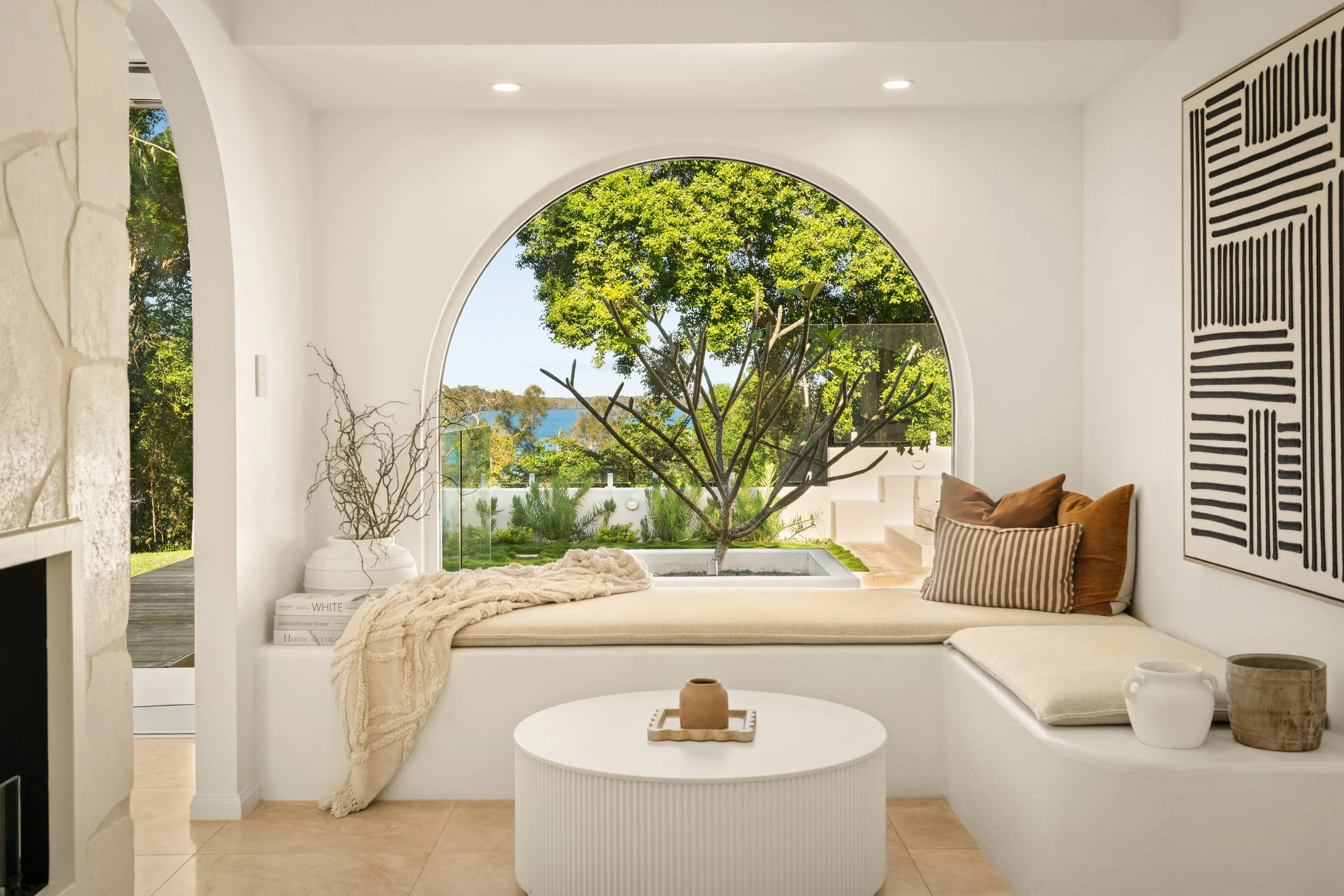 Cozy indoor space with a built-in window seat overlooking a lush garden and view of trees and water, with decorative pillows, a blanket, and a coffee table.