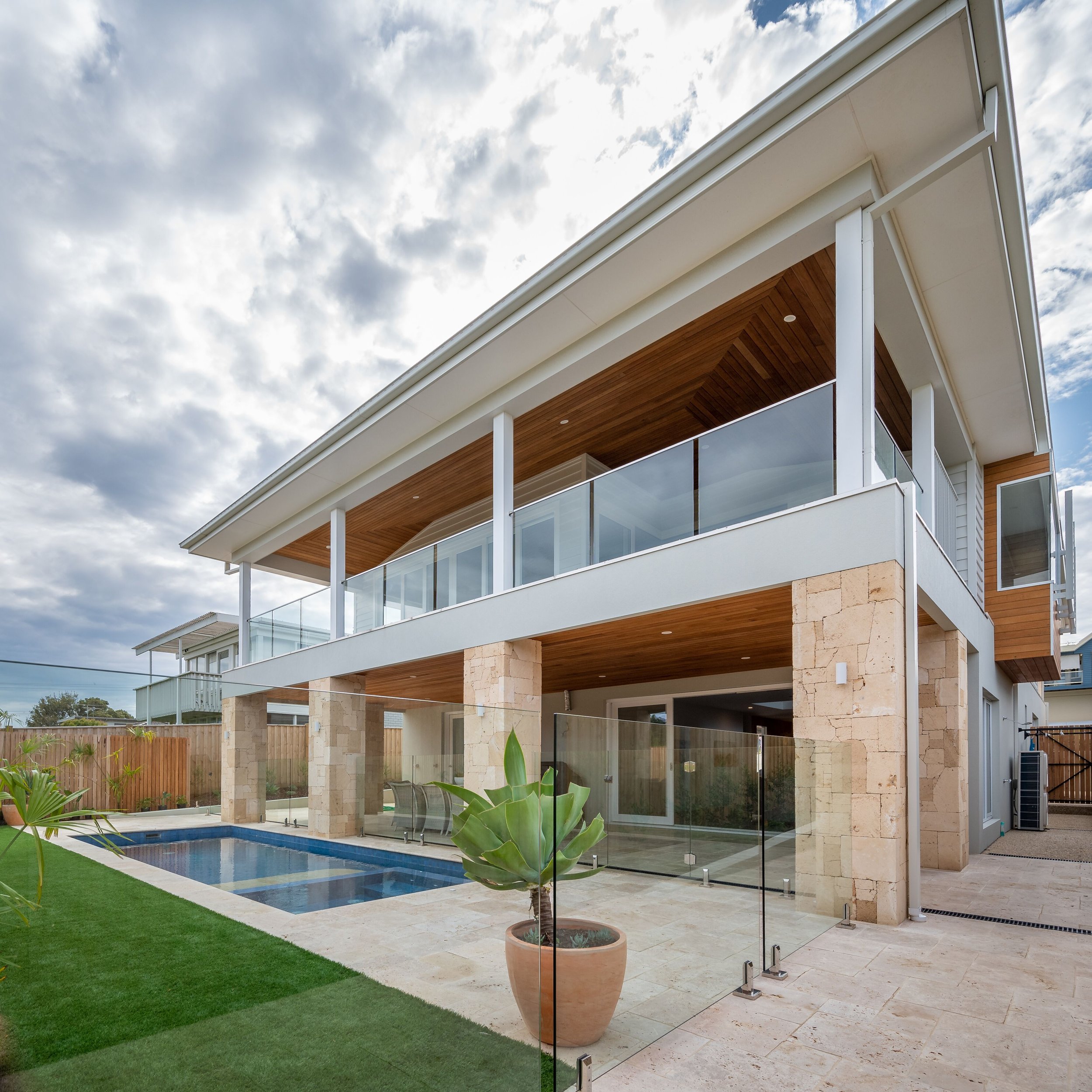 Modern multi-story house with glass balconies, a pool, grassy yard, and potted plants under a cloudy sky.