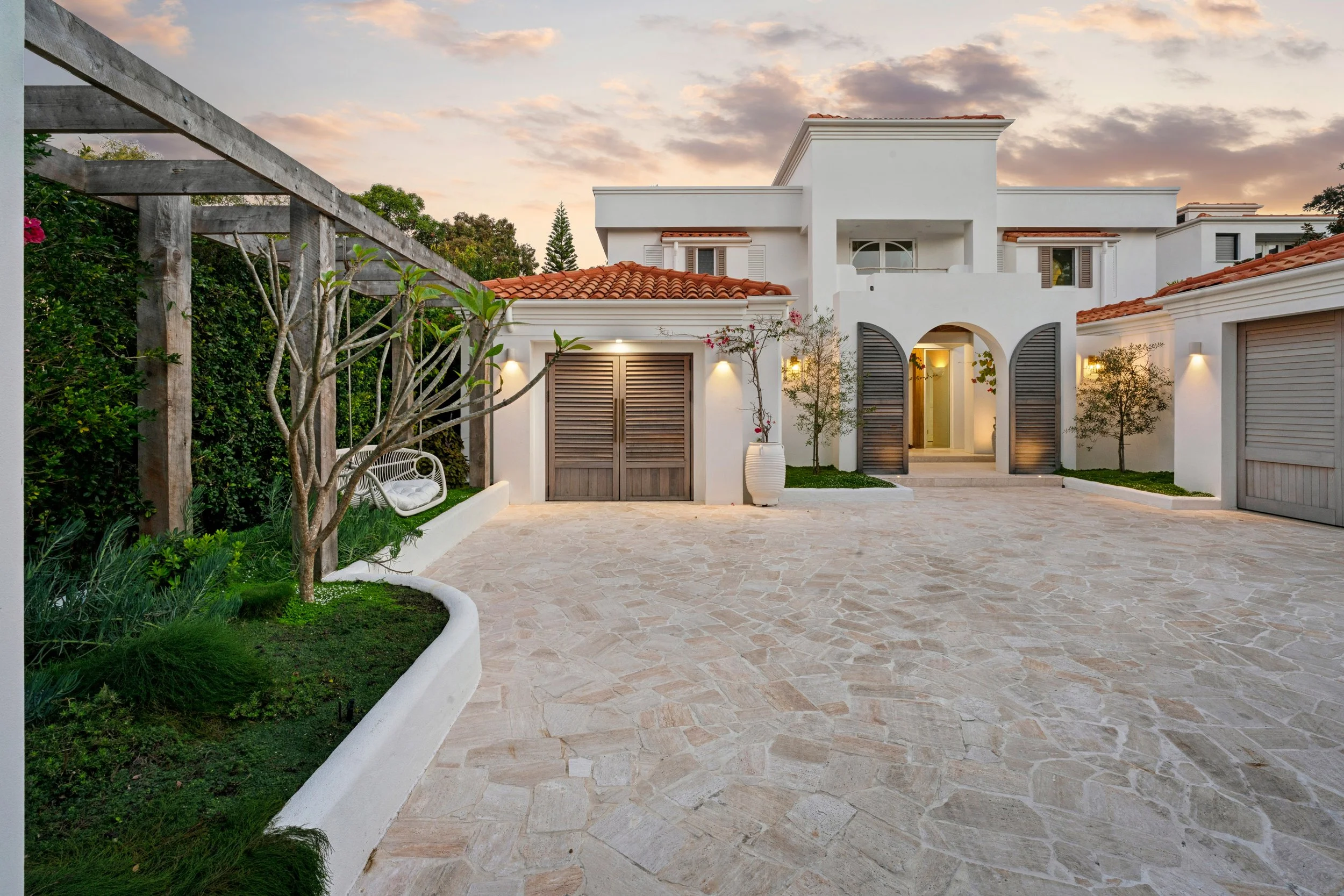 Luxury white house with red tile roof, surrounded by greenery, stone driveway, potted plants, and outdoor lighting at sunset.