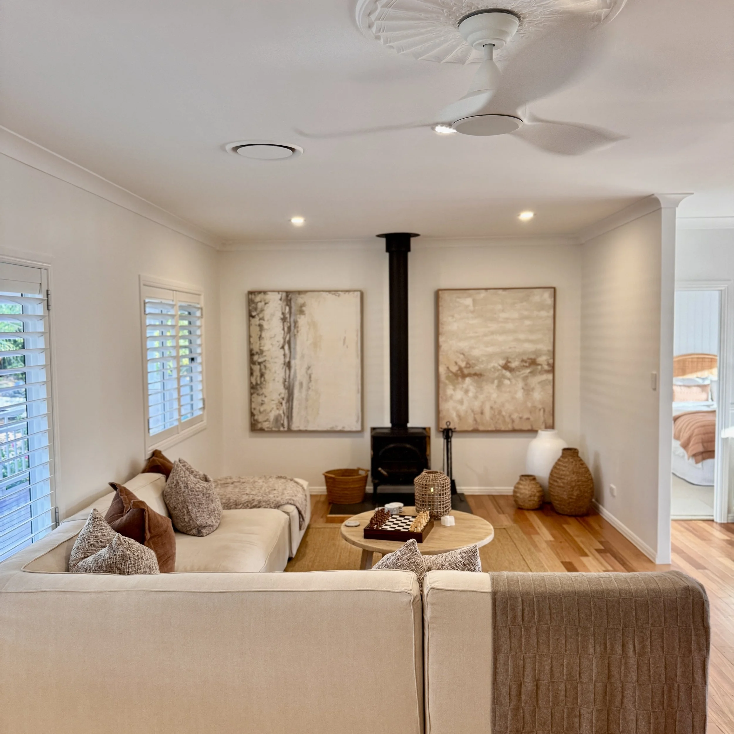 Living room with a white sectional sofa, pillows, a round wooden coffee table, a wood stove, abstract paintings, and decorative baskets in the corner.