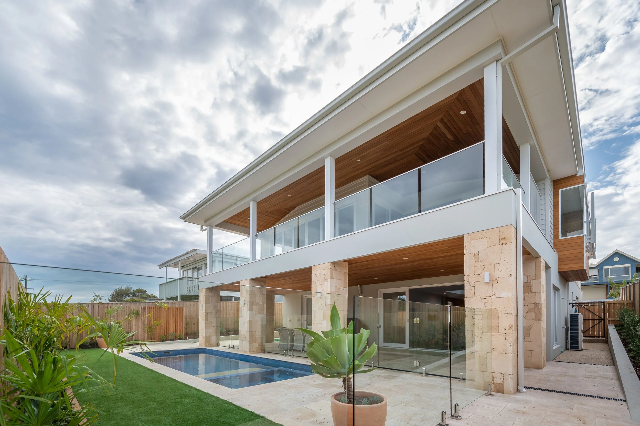 Modern two-story house with outdoor swimming pool, glass railing, wooden balcony ceiling, potted plant, and a grassy backyard, under a cloudy sky.