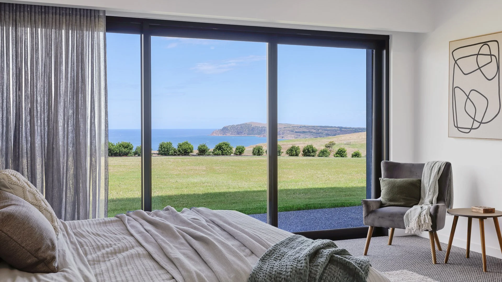 A bedroom with large glass window showing a view of green grass, bushes, a blue ocean, and a distant hill.