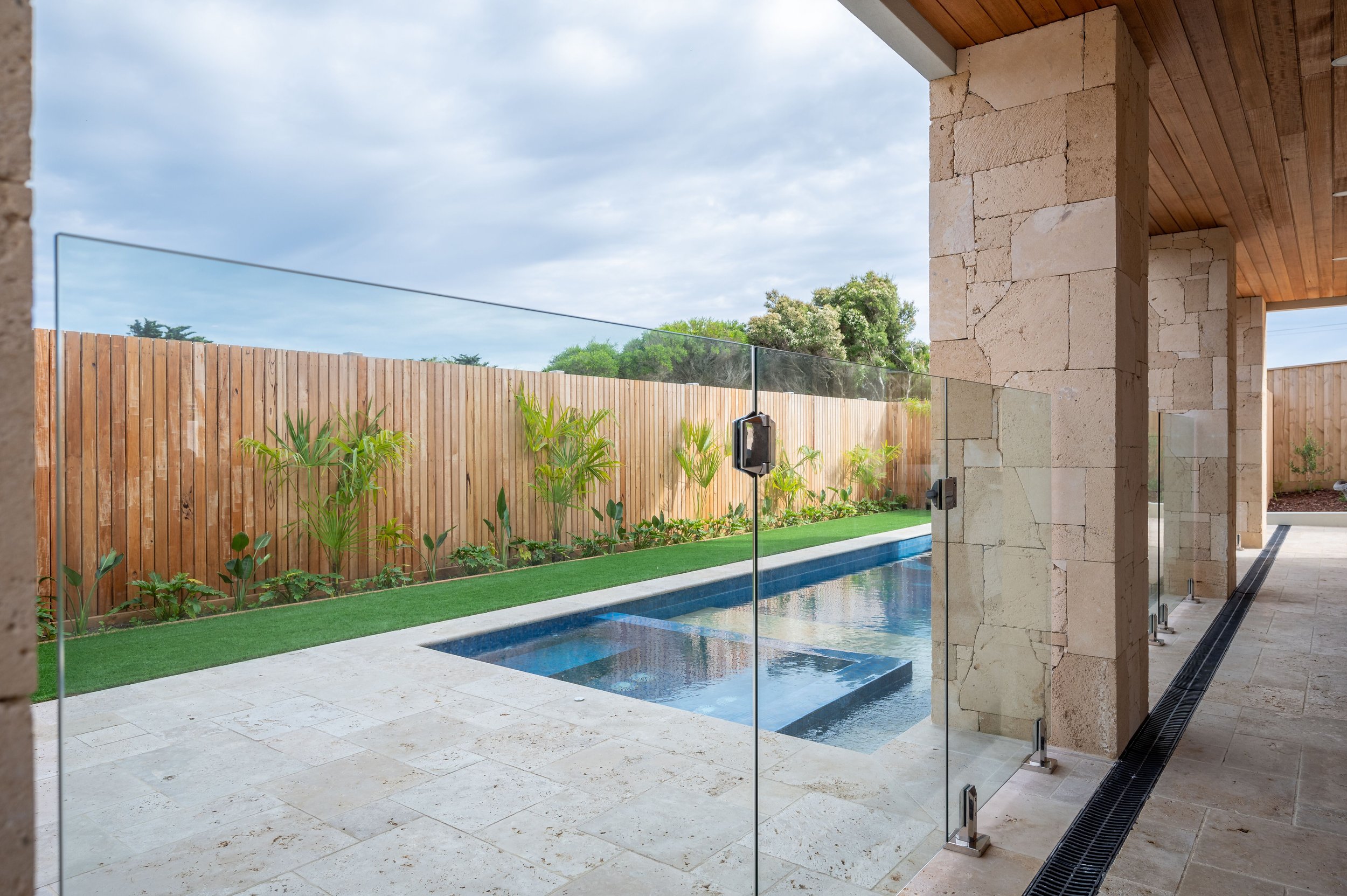 Modern backyard with a small rectangular pool, glass fencing, a wooden fence with plants, and stone columns supporting a wooden roof.