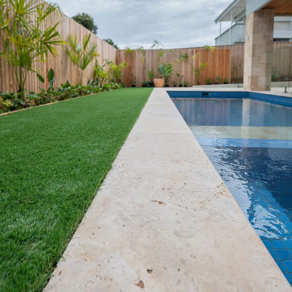 A backyard with a swimming pool, a concrete walkway, a green lawn, and a wooden fence with plants and potted foliage.