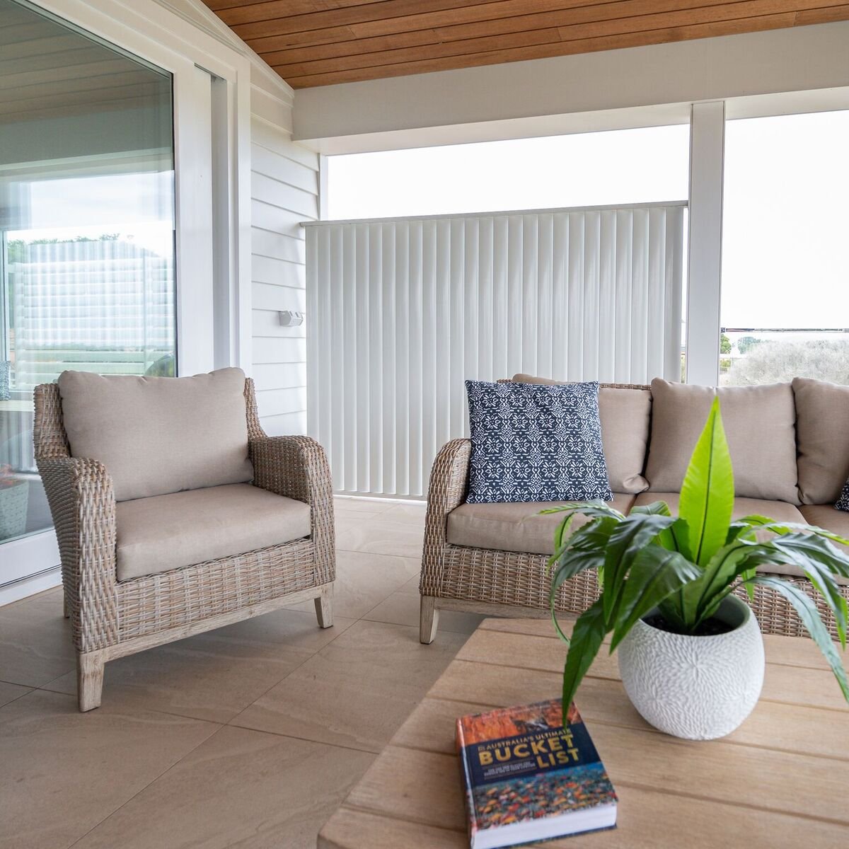 A cozy outdoor patio with wicker and cushioned seating, potted green plant, and a book on a wooden table, with a railing and sky in the background.