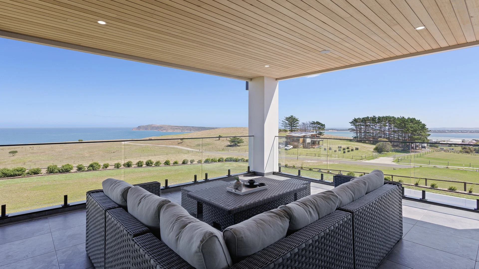 Balcony overlooking green lawn, trees, ocean, and clear blue sky, with wicker sofa and table