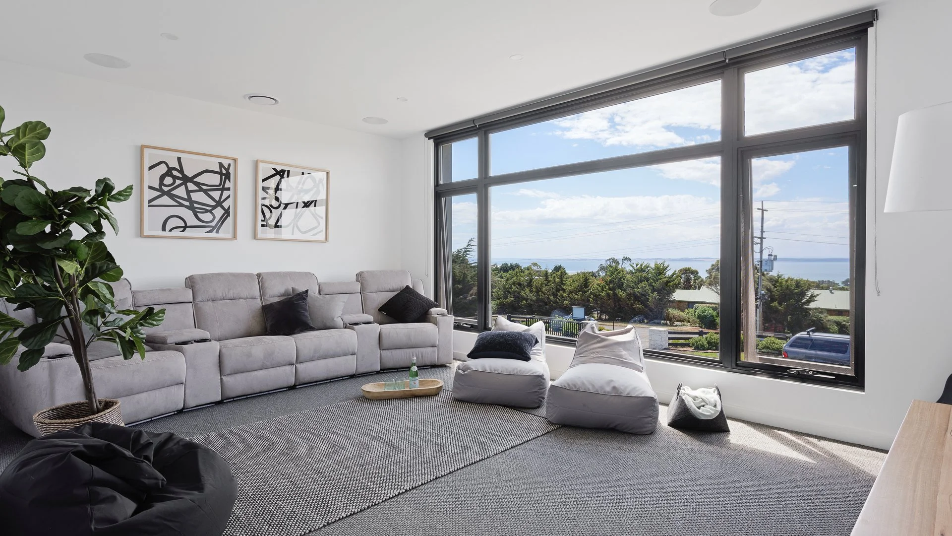 Living room with large windows showing a view of trees and water, light gray sofa with black pillows, abstract black and white wall art, potted plant, bean bag chairs, and a gray rug.