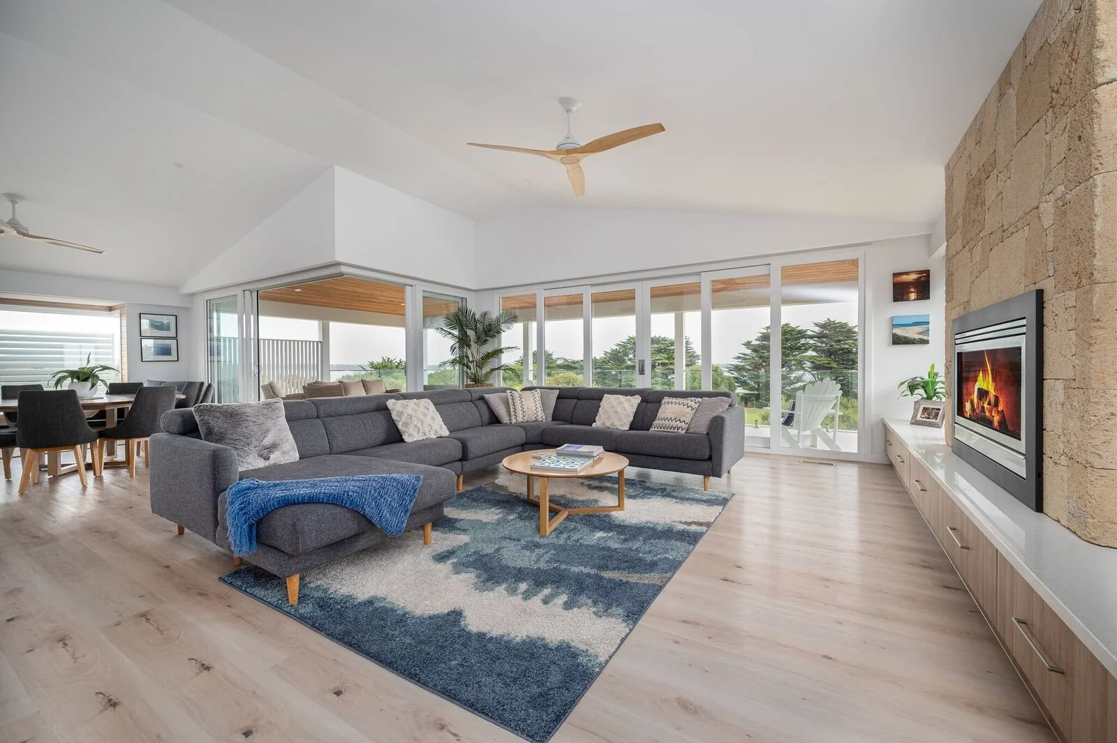 Living room with large gray sectional sofa, rug, coffee table, fireplace, and sliding glass doors opening to outdoor patio with view of trees.