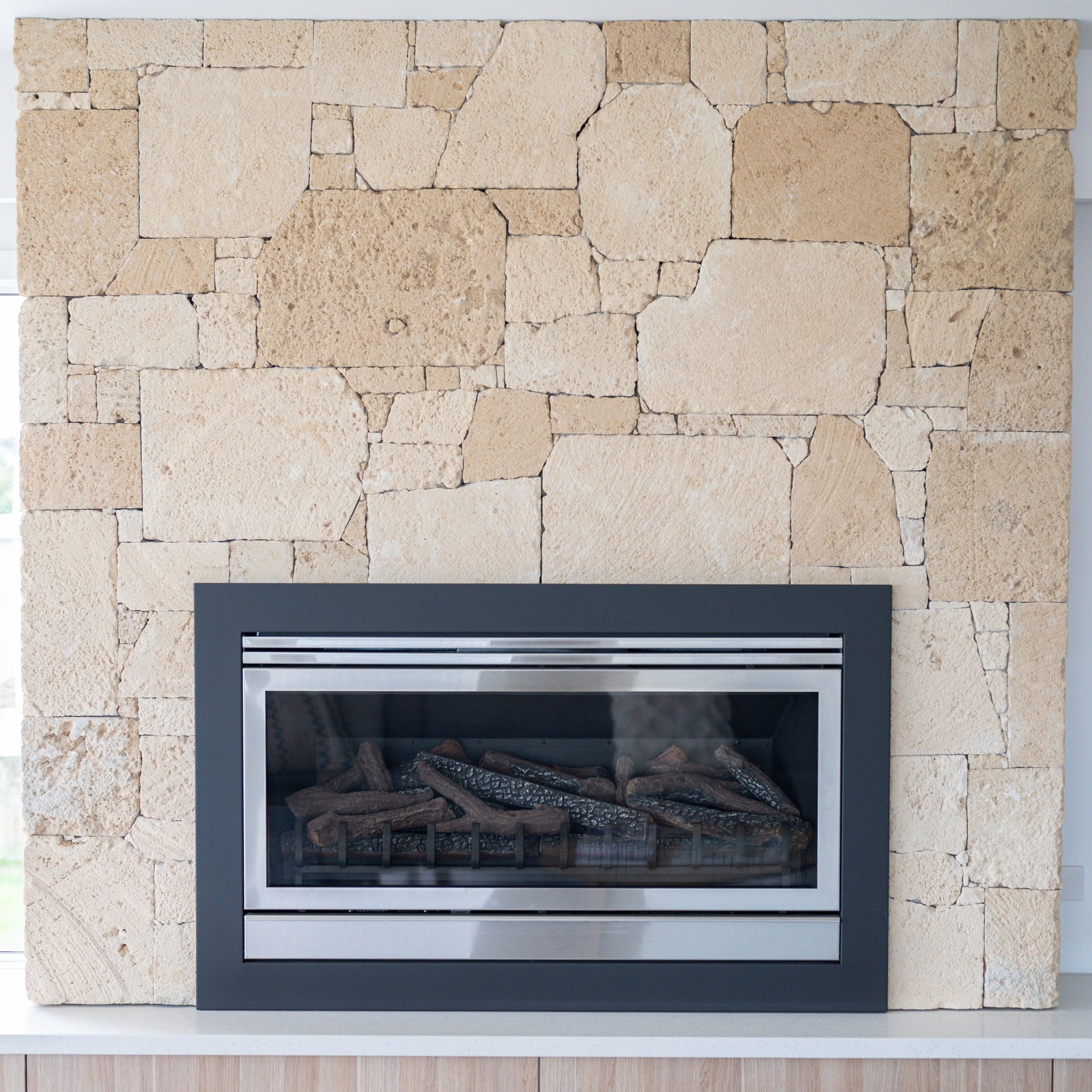 Modern fireplace set in a beige stone wall with gas logs inside the glass enclosure.