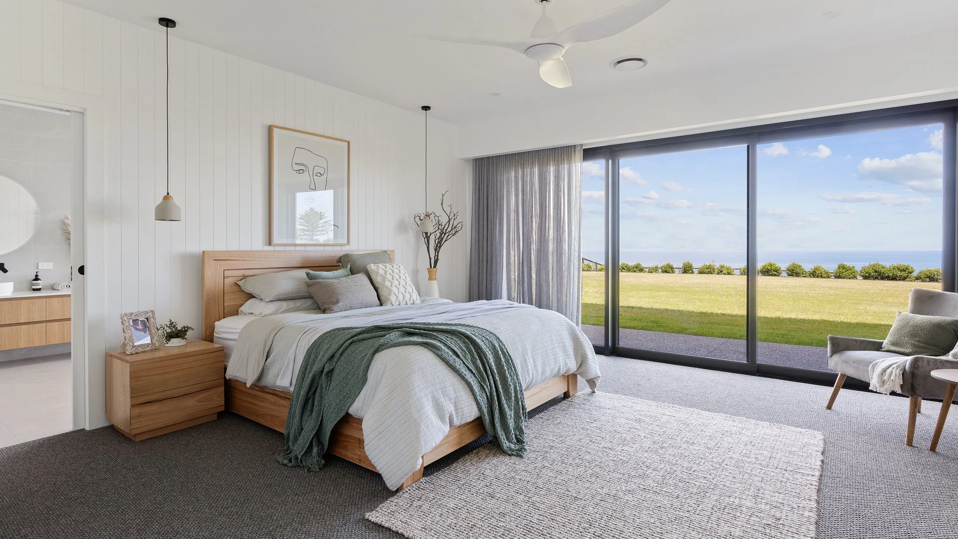Modern bedroom with large sliding glass doors overlooking a grassy area and ocean, featuring a wooden bed with neutral bedding, a bedside table with a picture frame and plant, and a cozy chair with a pillow near the window.