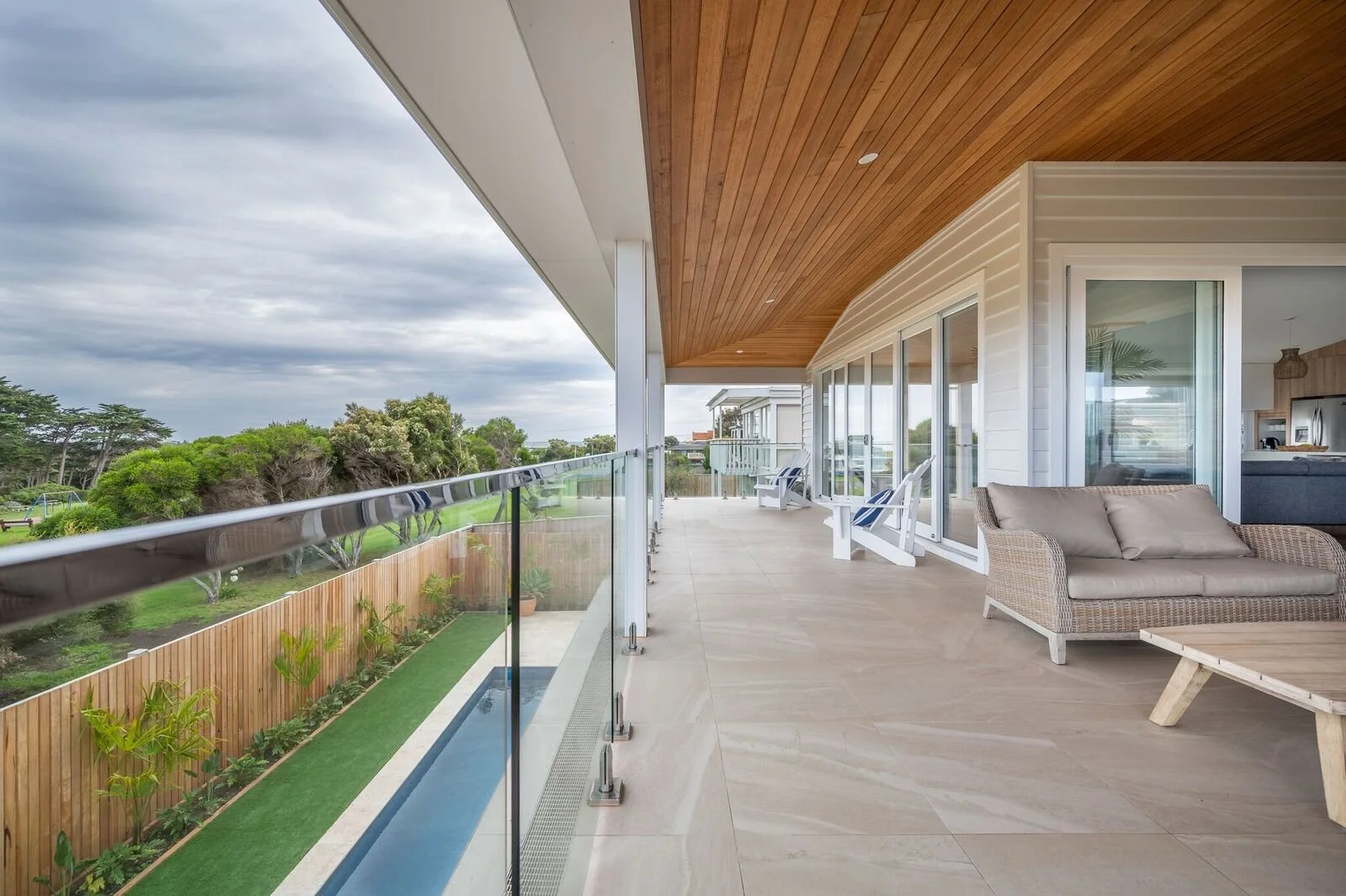 Spacious balcony with wicker sofa, white chairs with blue cushions, and a glass railing overlooking a grassy yard with trees and a cloudy sky.