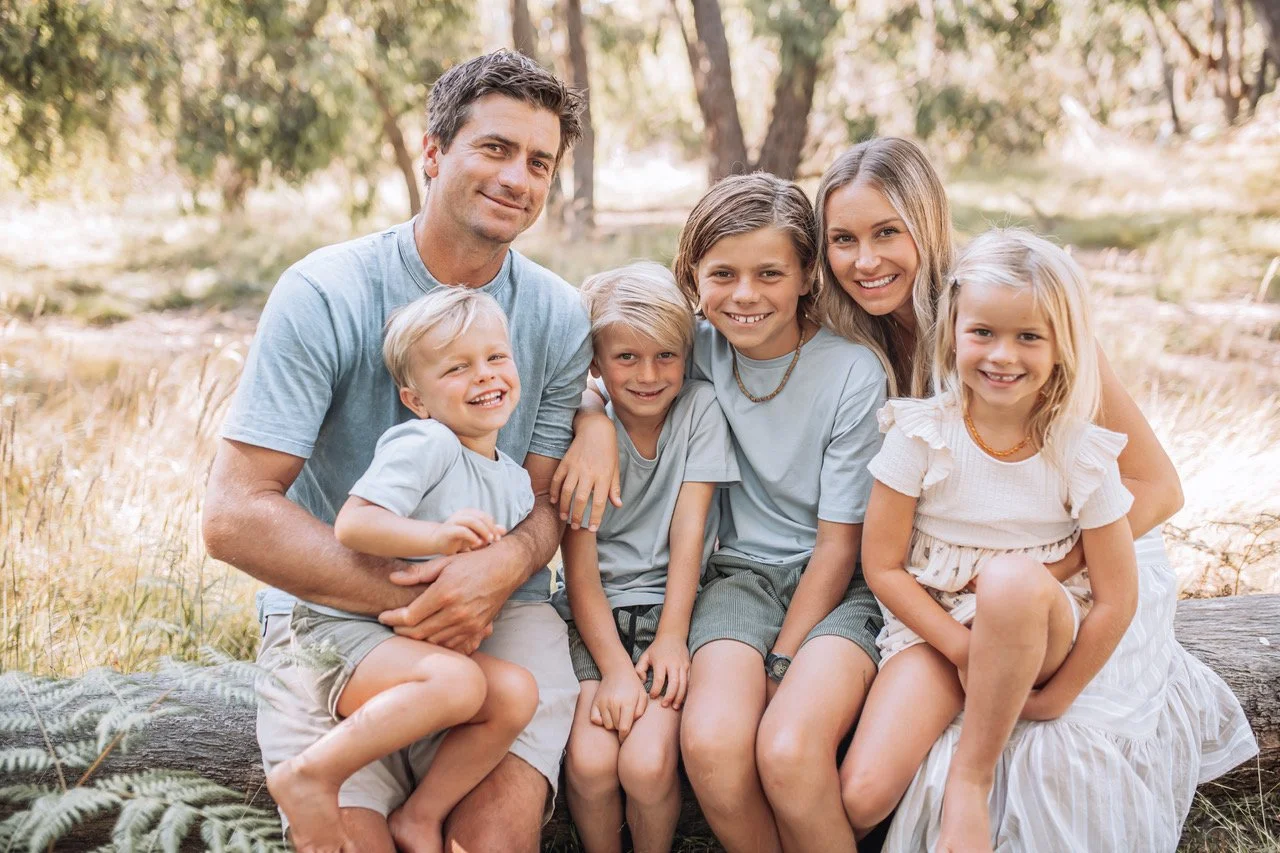 A family of six smiling and sitting together on a log outdoors in a forested area during daytime.