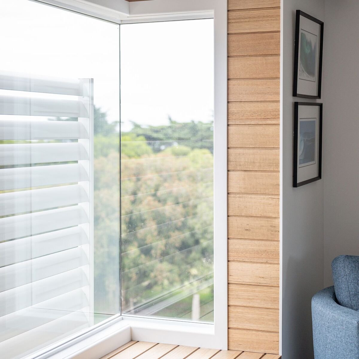 Corner of a room with a large glass window, white window shutters, a wood-paneled wall, and two framed pictures on a white wall.