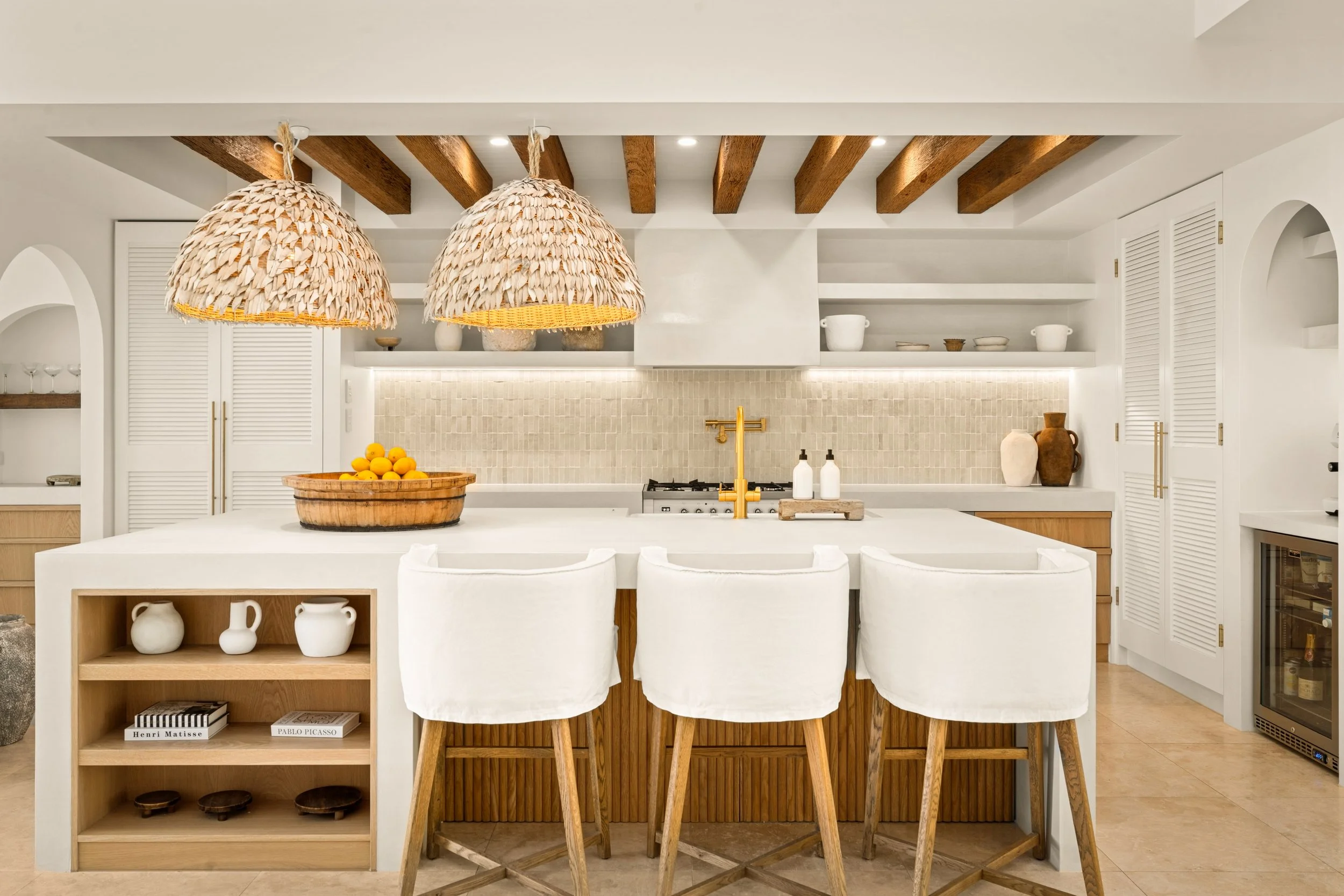 Modern kitchen with white island counter, four white chairs, hanging woven pendant lights, wood beams ceiling, open shelving, beige backsplash, and decorative pottery.