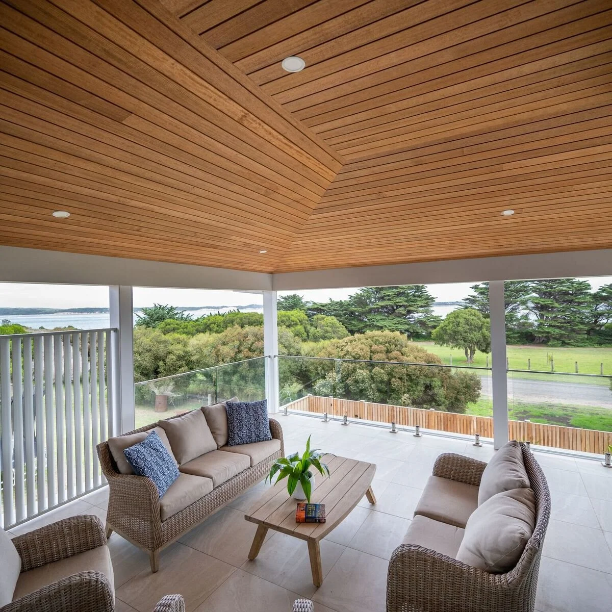 Covered outdoor balcony with wicker sofas, wooden coffee table, view of trees and water, wooden ceiling, glass railing.
