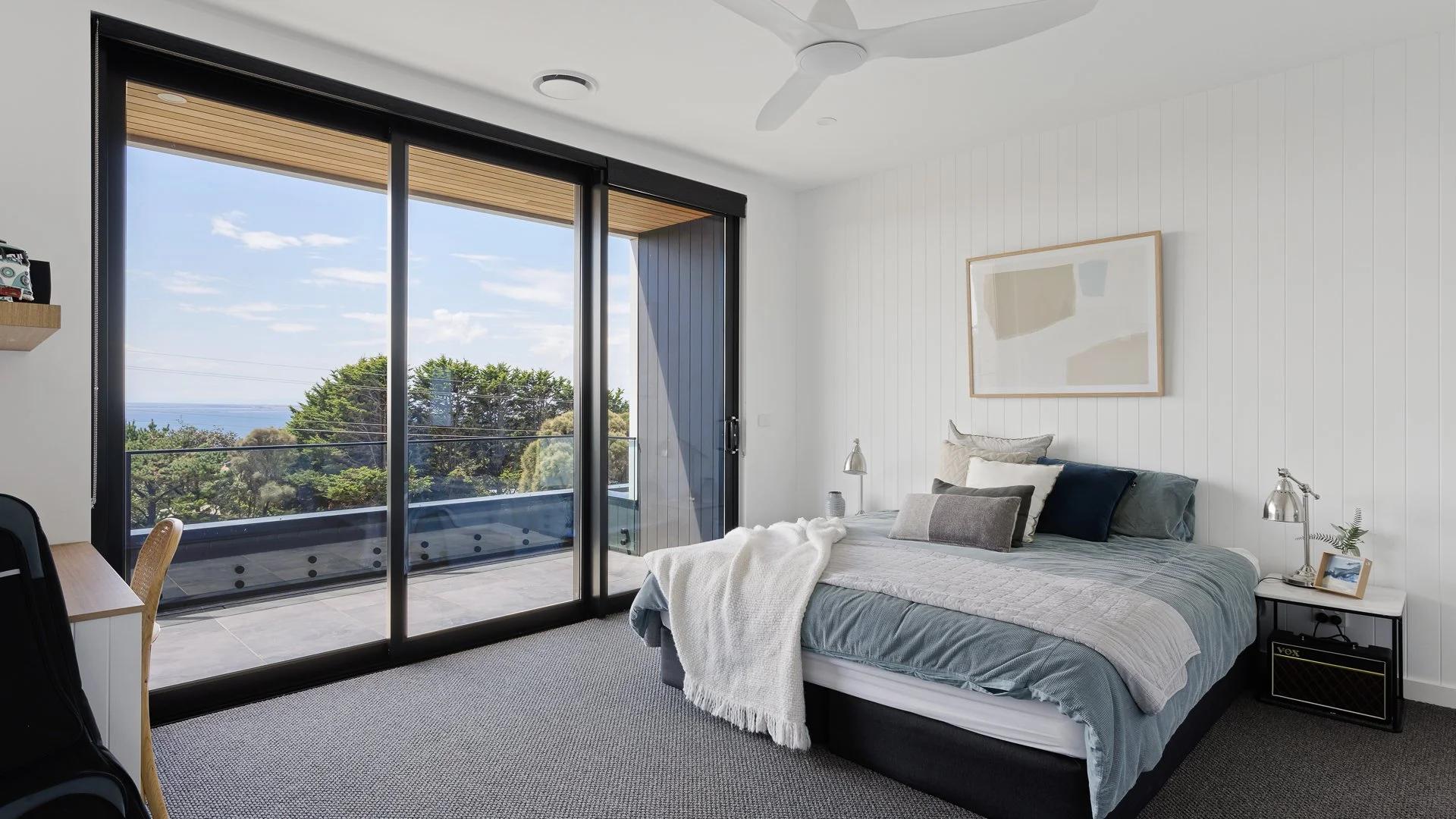 Bedroom with a large sliding glass door leading to a balcony, with a view of trees and a body of water in the distance. The room has a bed with gray and white bedding, pillows, a white throw blanket, and a nightstand with a lamp, picture frame, and s