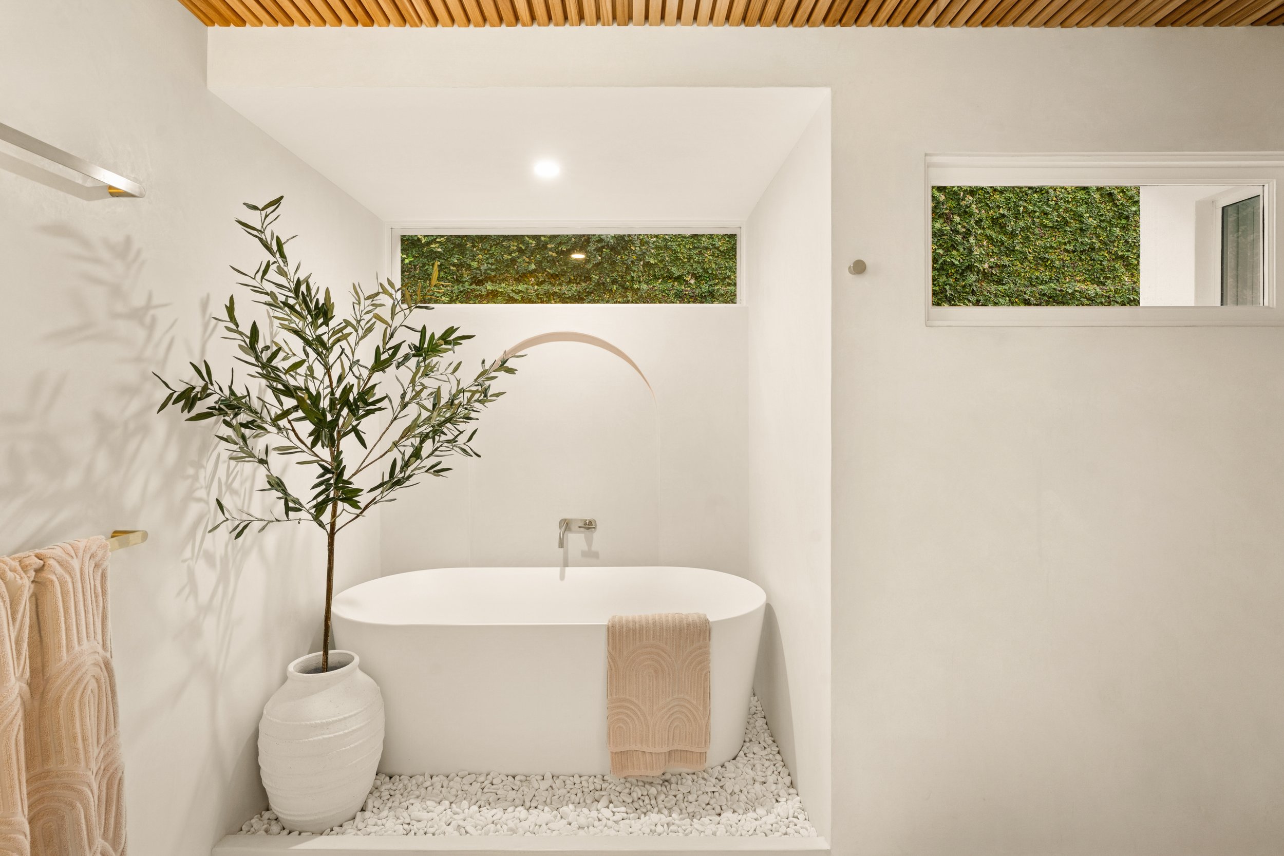 A modern minimalist bathroom with a white bathtub, a large potted plant, and two narrow horizontal windows with greenery outside.