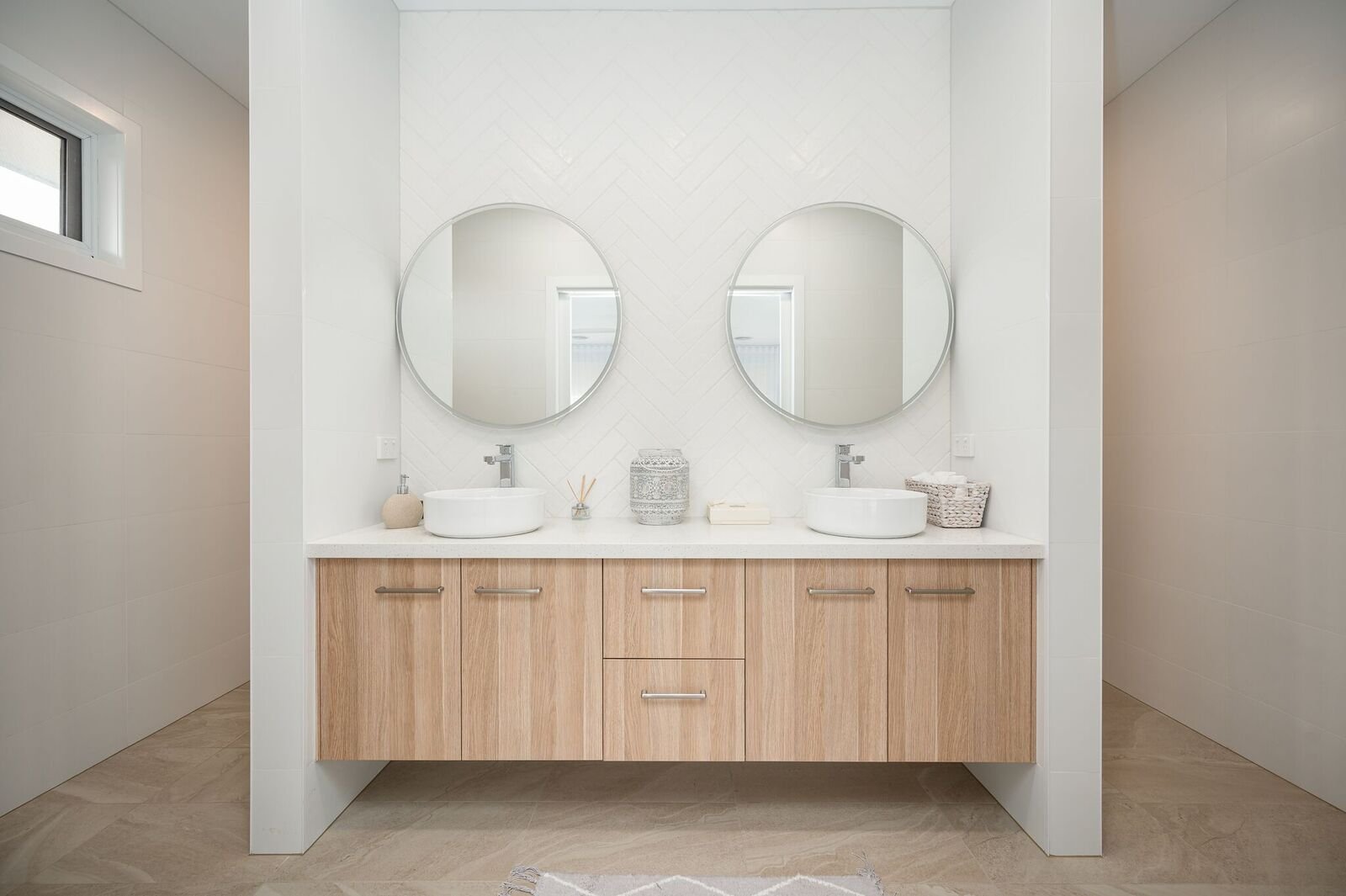 Modern bathroom with two white vessel sinks and round mirrors on a white wall. Wooden vanity with six drawers and two cabinets, small decor items and a basket on countertop. Light-colored tile floor and walls, small window on the left.