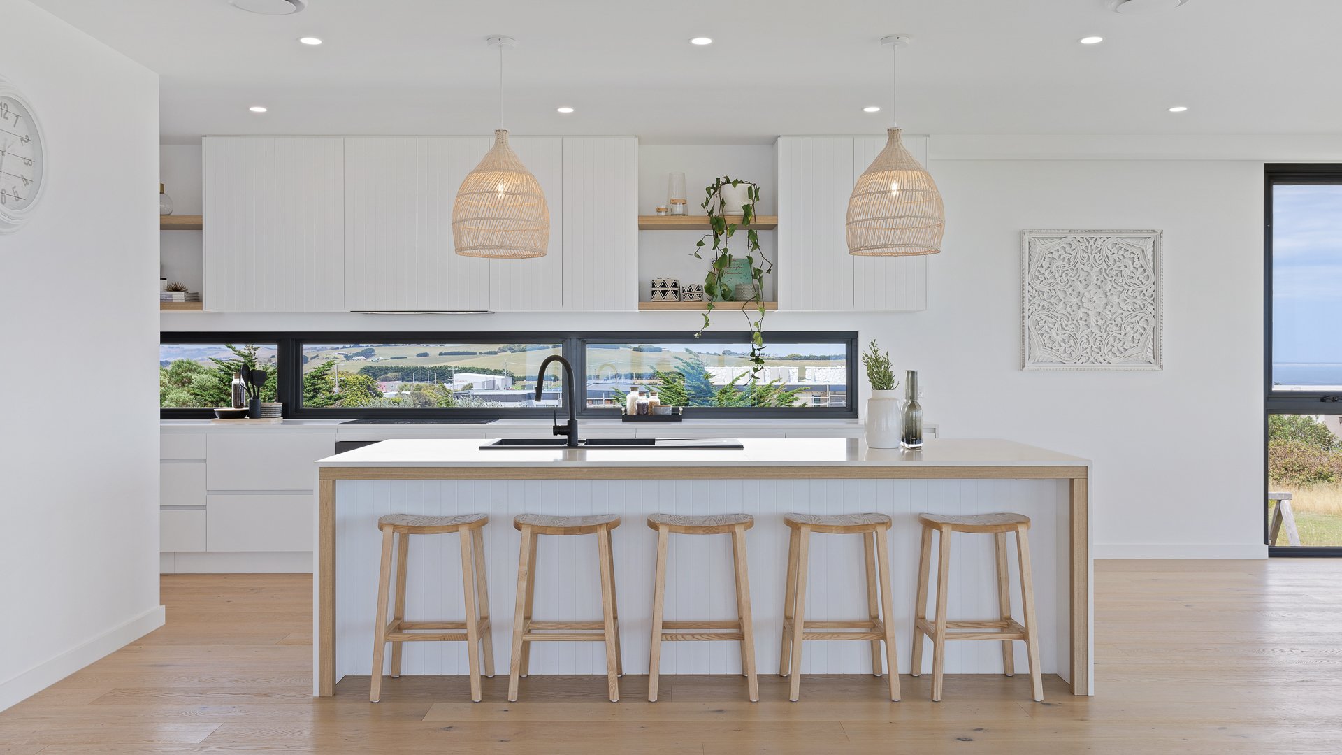 Modern white kitchen with island, wooden stools, black faucet, and large windows showing outdoor landscape