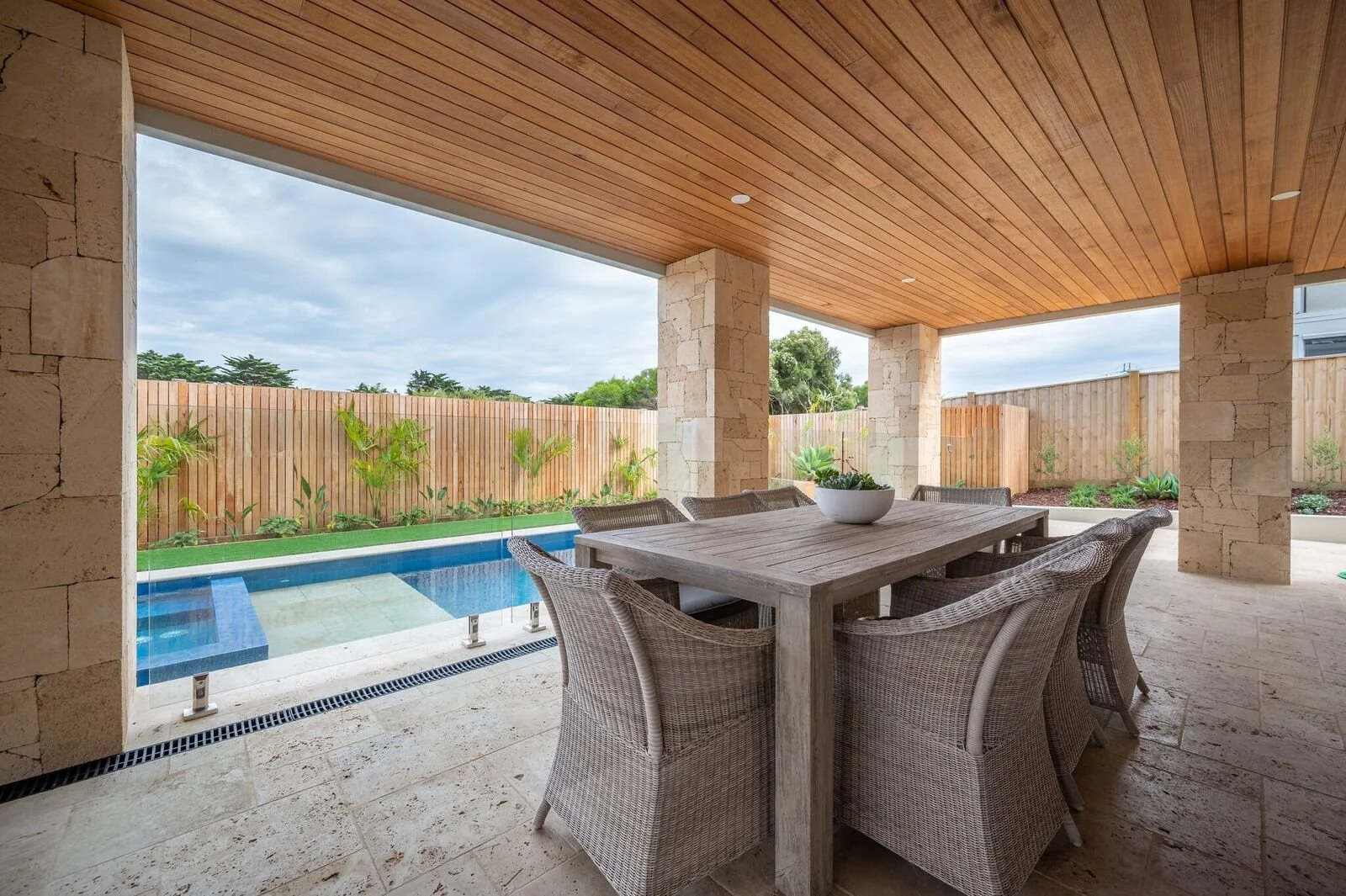 Covered outdoor patio with a wooden ceiling, stone pillars, a rectangular dining table with woven chairs, and a backyard pool enclosed by a wooden fence with tropical plants.