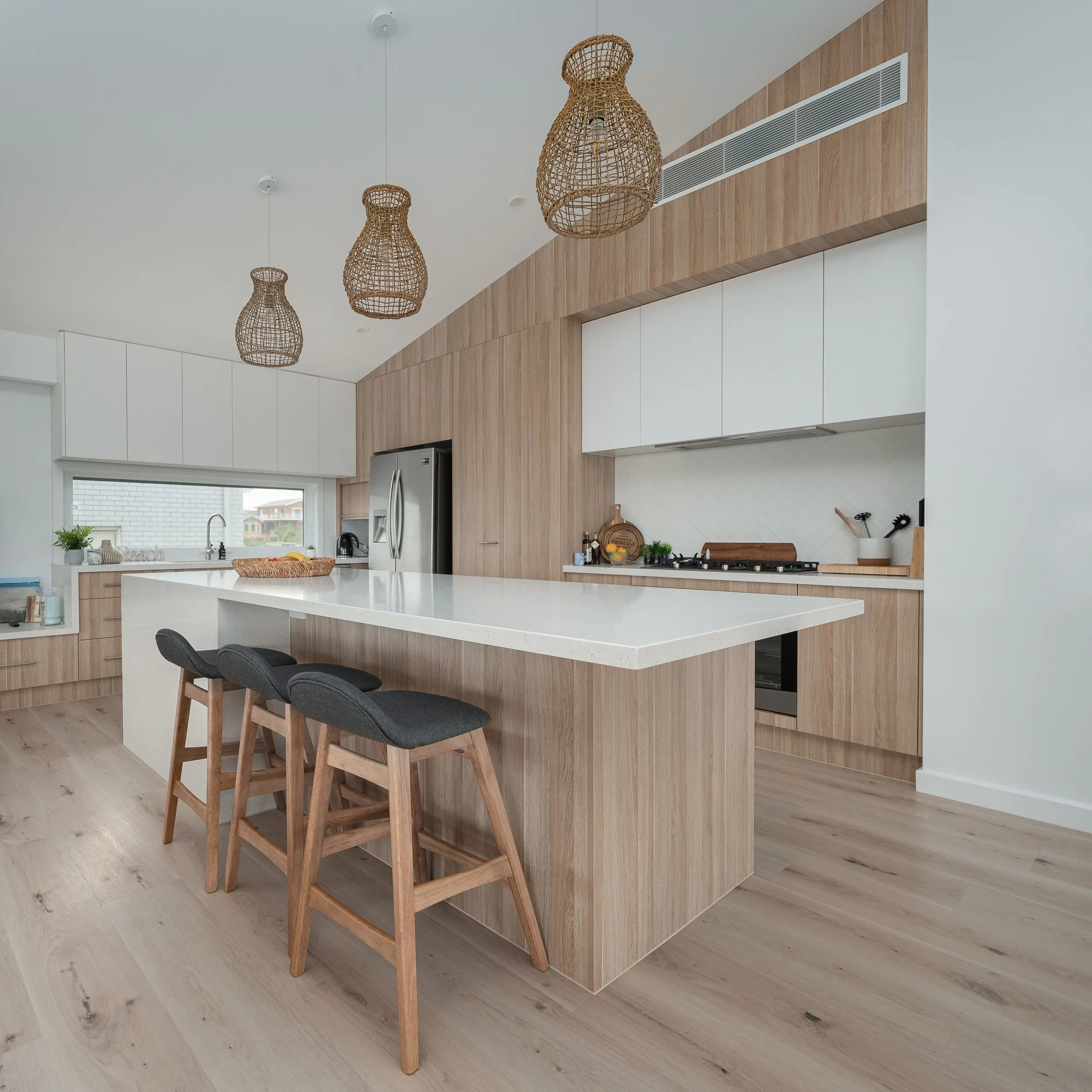 Modern kitchen with white countertops, light wood cabinetry, stainless steel refrigerator, and black bar stools with fabric seats.