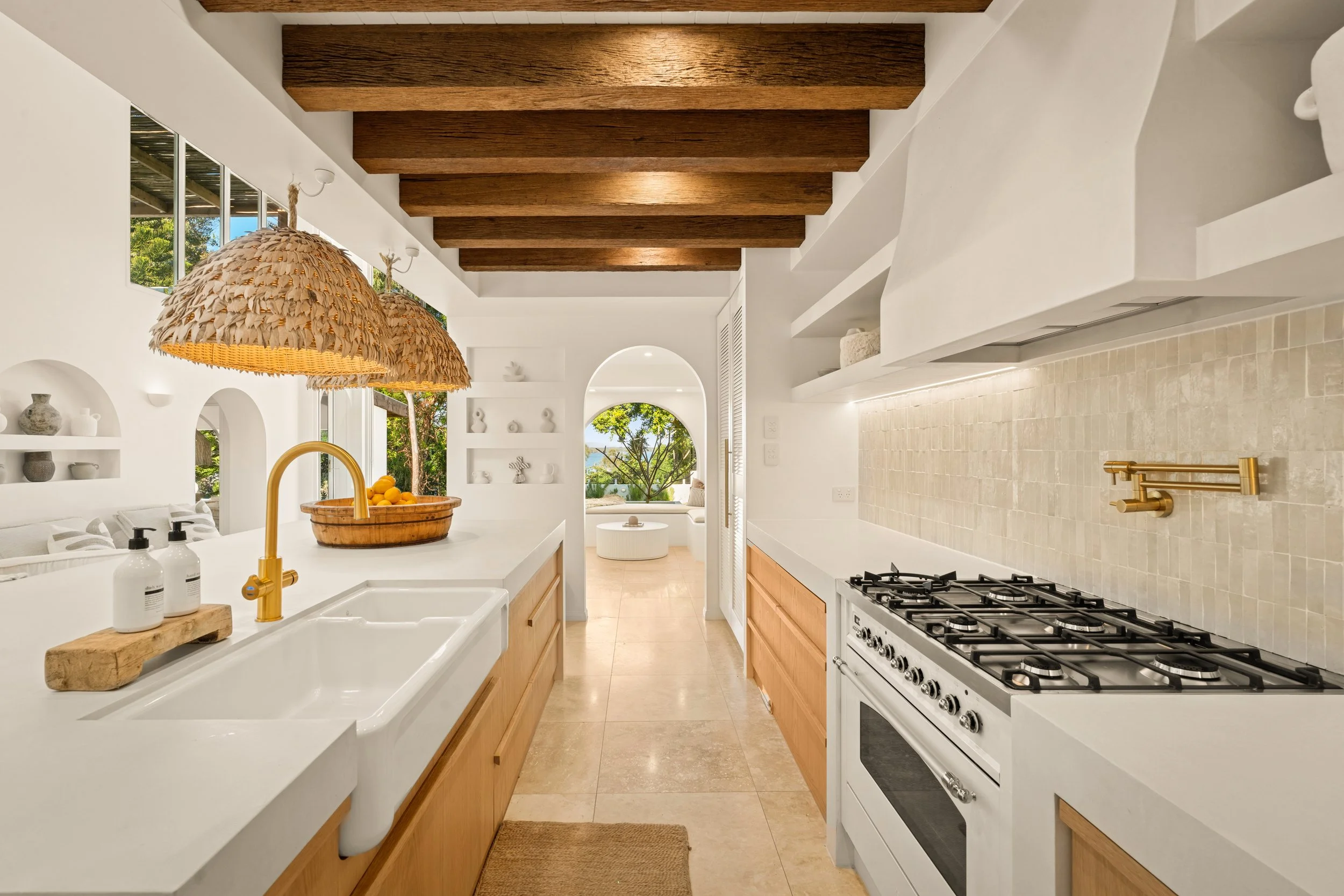 Bright kitchen with white countertops, wooden cabinets, beige tiled backsplash, gas stove, and a large woven pendant light. Open living space with large windows and a view of trees outside.