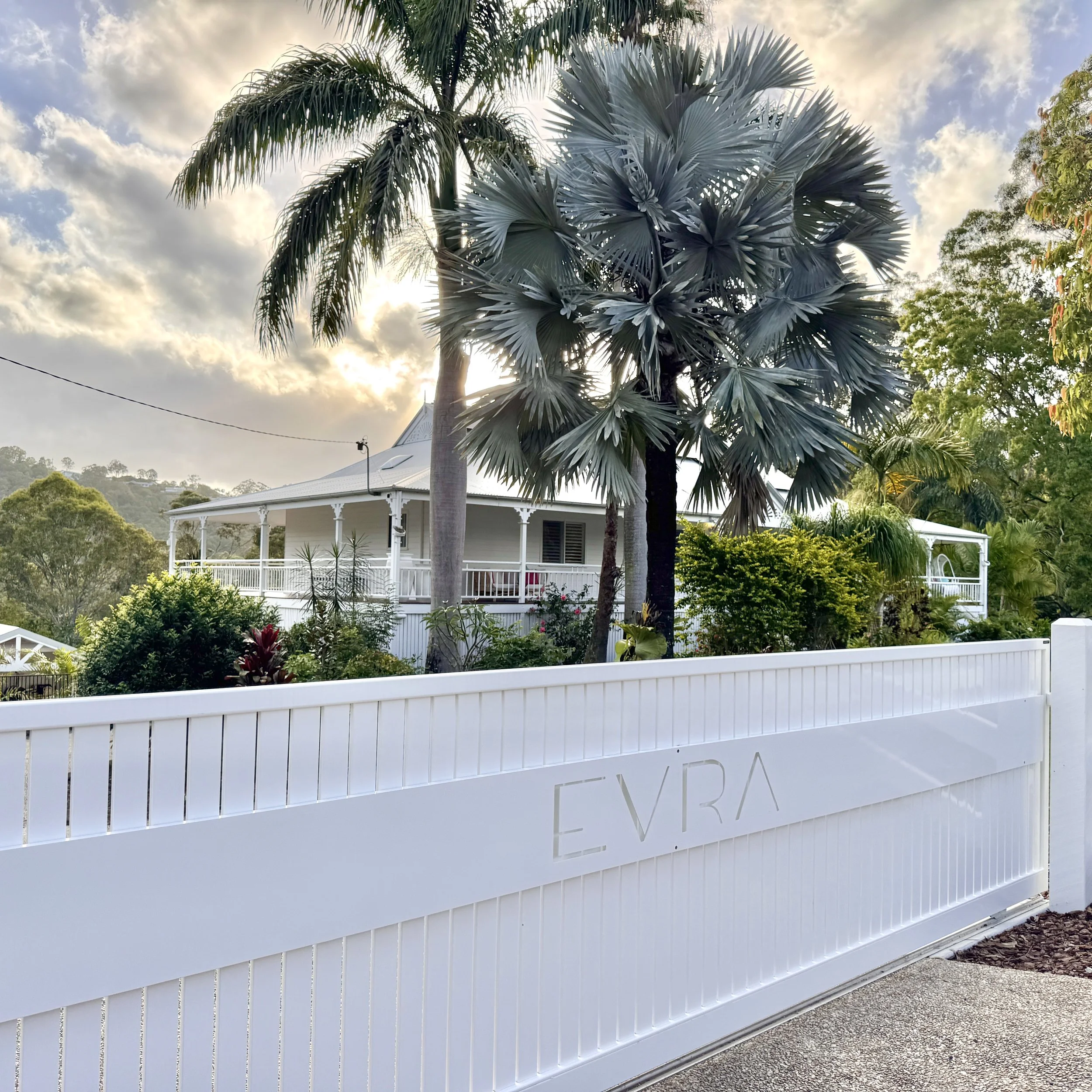 White gate with the word 'EVRA' in front of a large house with tropical trees and green foliage, under a partly cloudy sky.