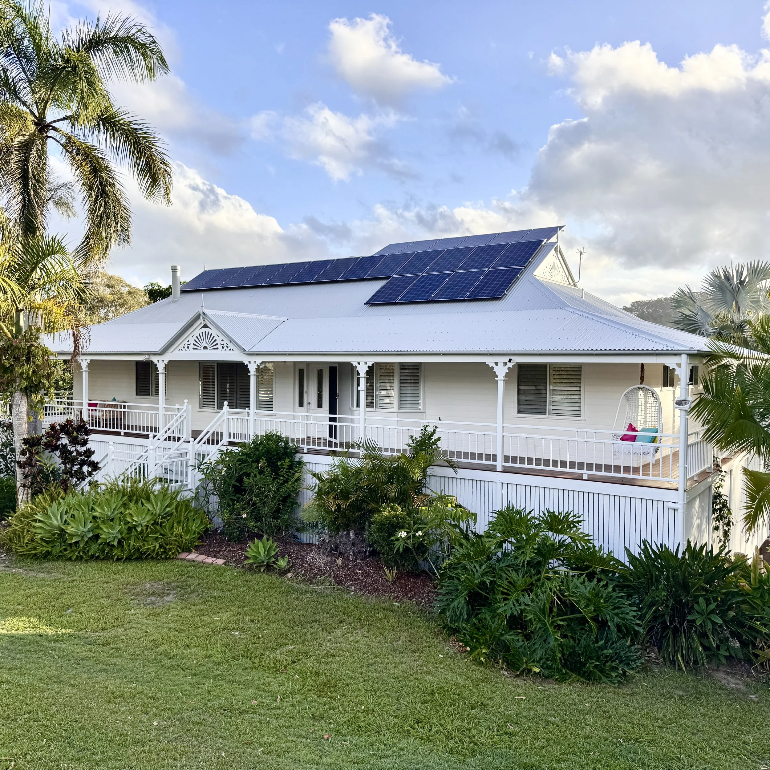 A white, two-story house with a metal roof and solar panels, surrounded by lush green plants and palm trees.