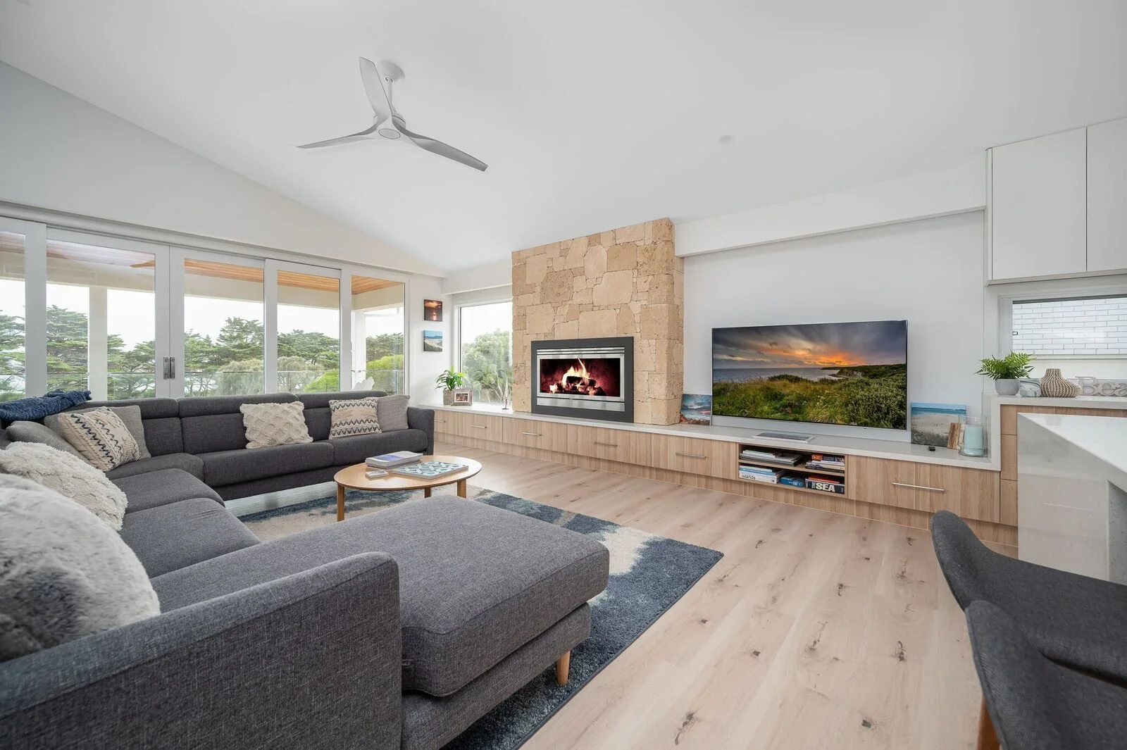Modern living room with large gray sectional sofa, fireplace, flat-screen TV, and floor-to-ceiling windows overlooking greenery.