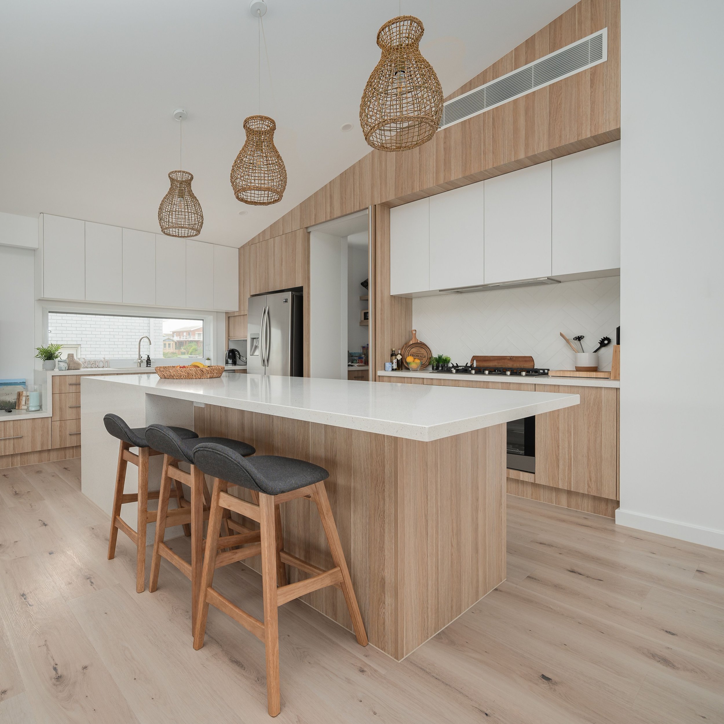 Modern kitchen with a large white island, three gray-barstool chairs, light wood cabinetry, stainless steel appliances, woven pendant lights, and a window overlooking houses.