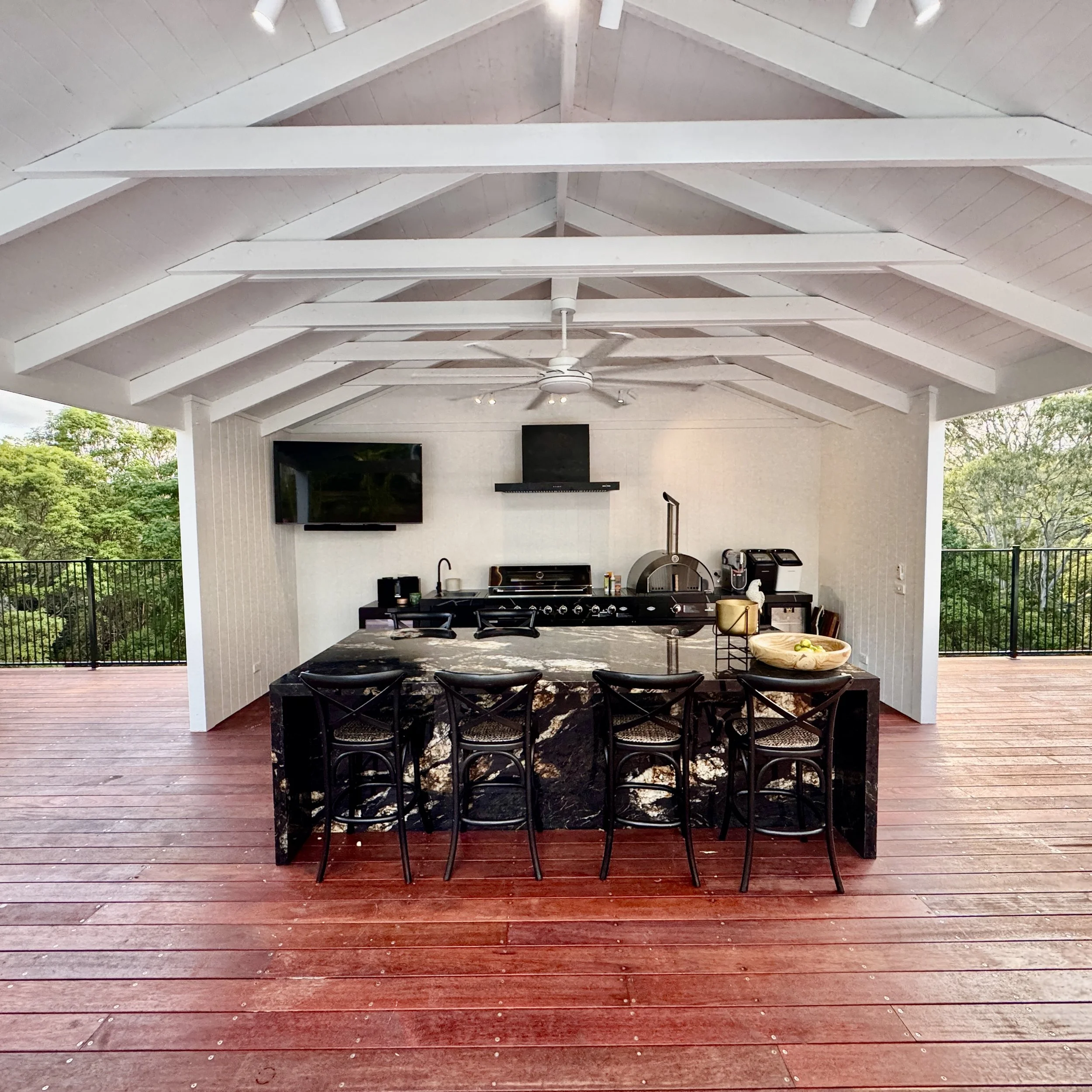 Outdoor kitchen area with a black marble island, six black bar stools, a ceiling fan, mounted TV, and various appliances, on a wooden deck with trees in the background.