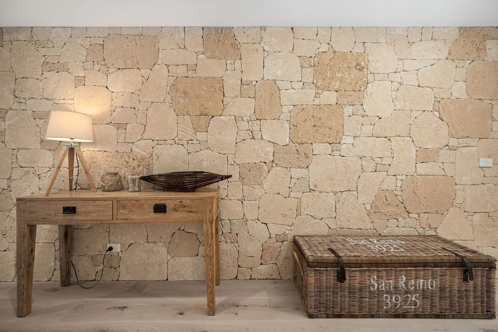 Room with a stone wall, a wooden table with a lamp and decorative items, and a wicker storage chest on a light-colored wooden floor.