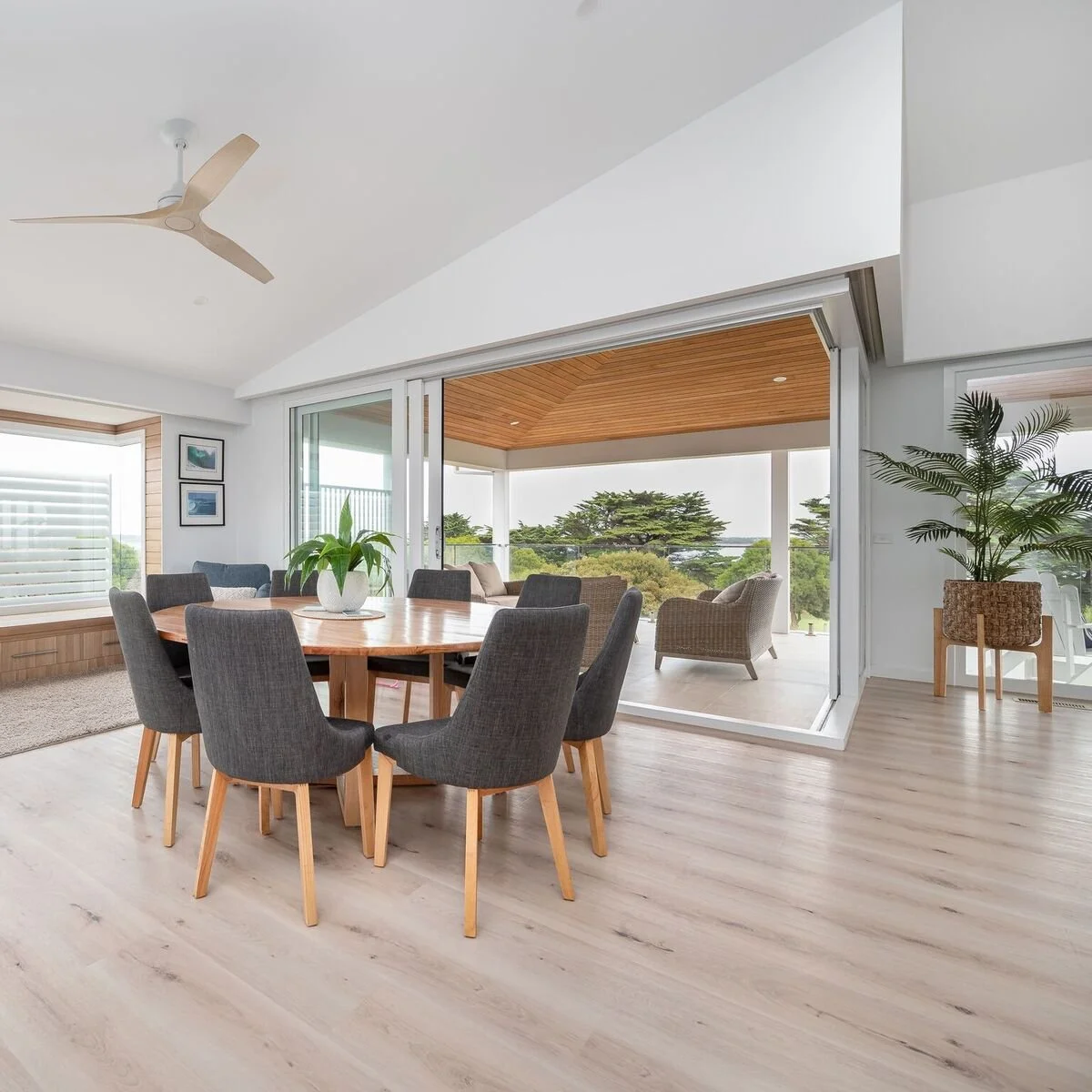 Modern dining room with hardwood floor, six gray upholstered chairs around a round wooden table with a potted plant centerpiece, large sliding glass doors leading to a balcony with outdoor seating, and large windows with natural light and views of tr