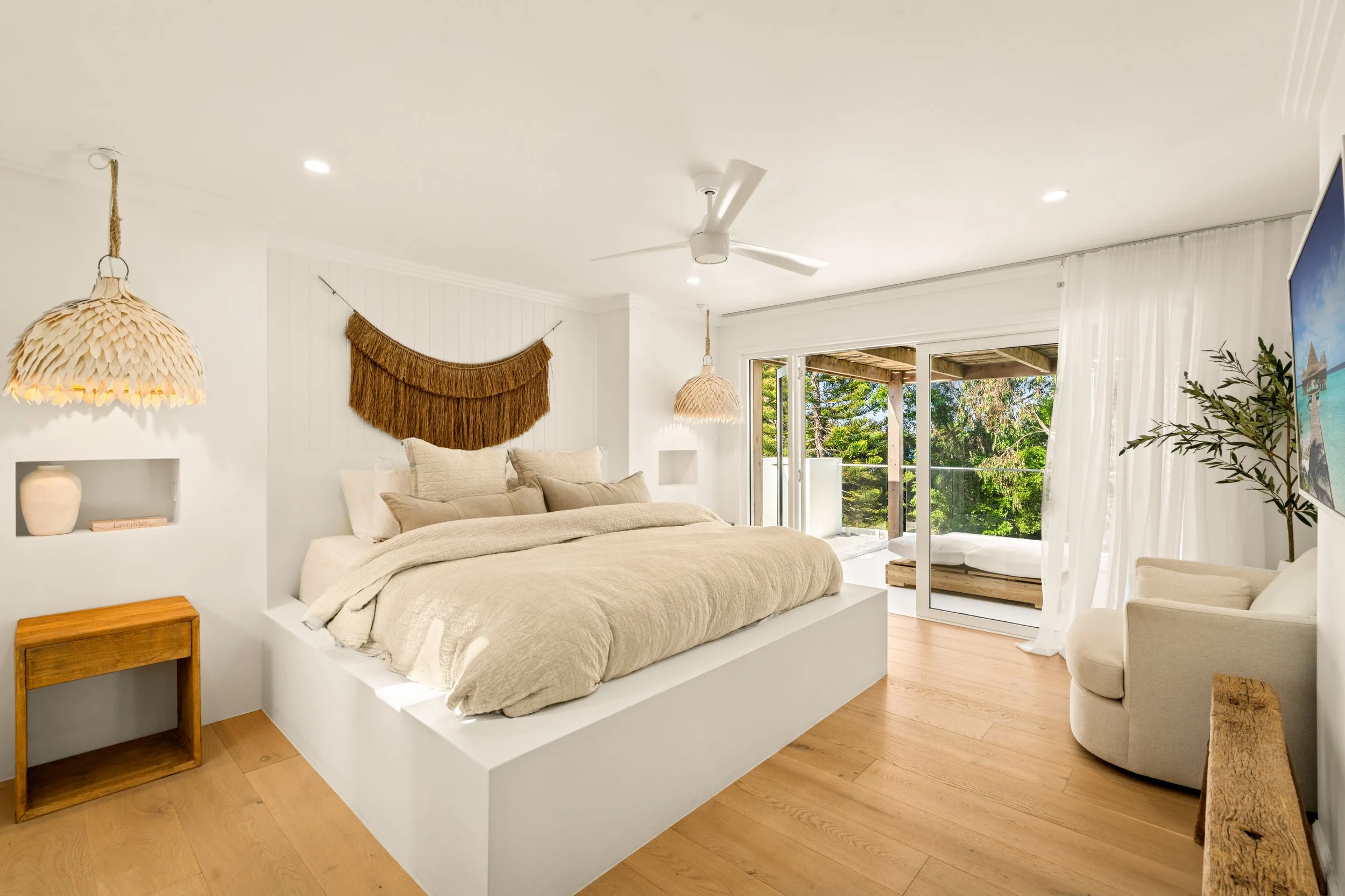 Bright bedroom with a large bed, beige bedding, and decorative pillows. Wooden nightstand, woven pendant lights, wall hanging art, and sliding glass door opening to a balcony with trees outside.