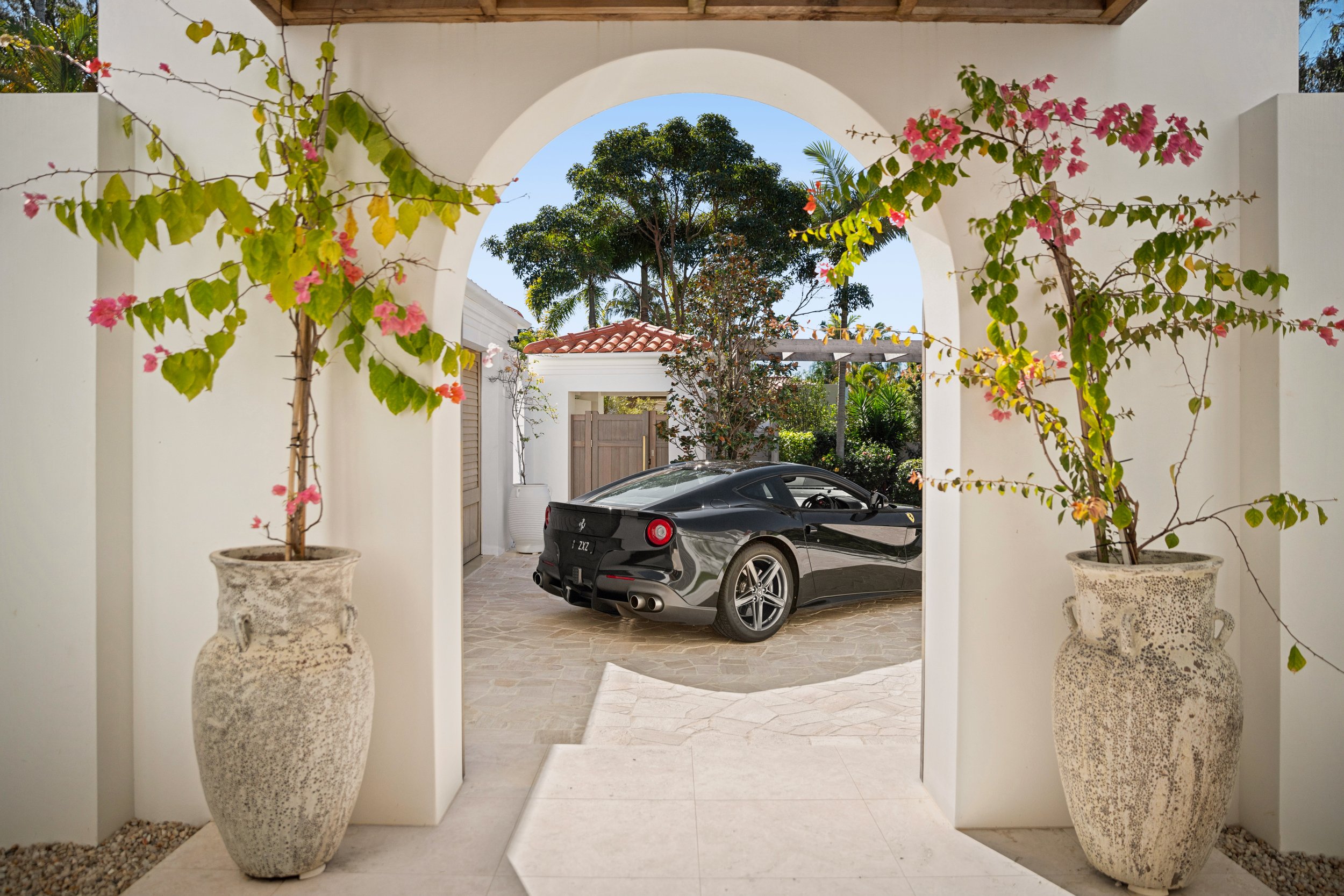 A view through an archway with flowering plants in large decorative pots, revealing a black sports car parked in a Mediterranean-style courtyard with trees and a red-tiled roof building in the background.