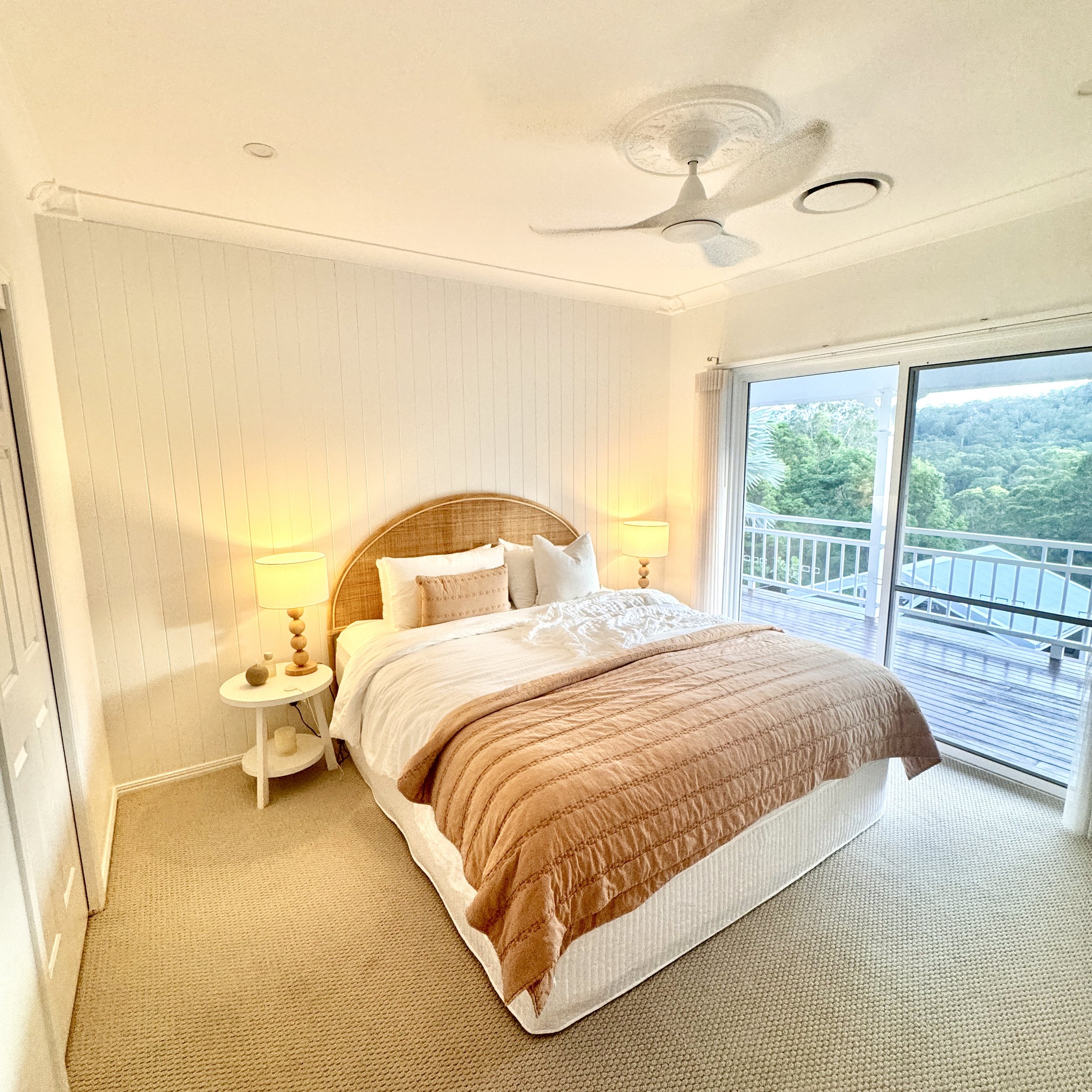 A bedroom with a large bed, beige bedding, and a wicker headboard, two bedside lamps, a sliding glass door leading to a balcony with a view of trees and hills.