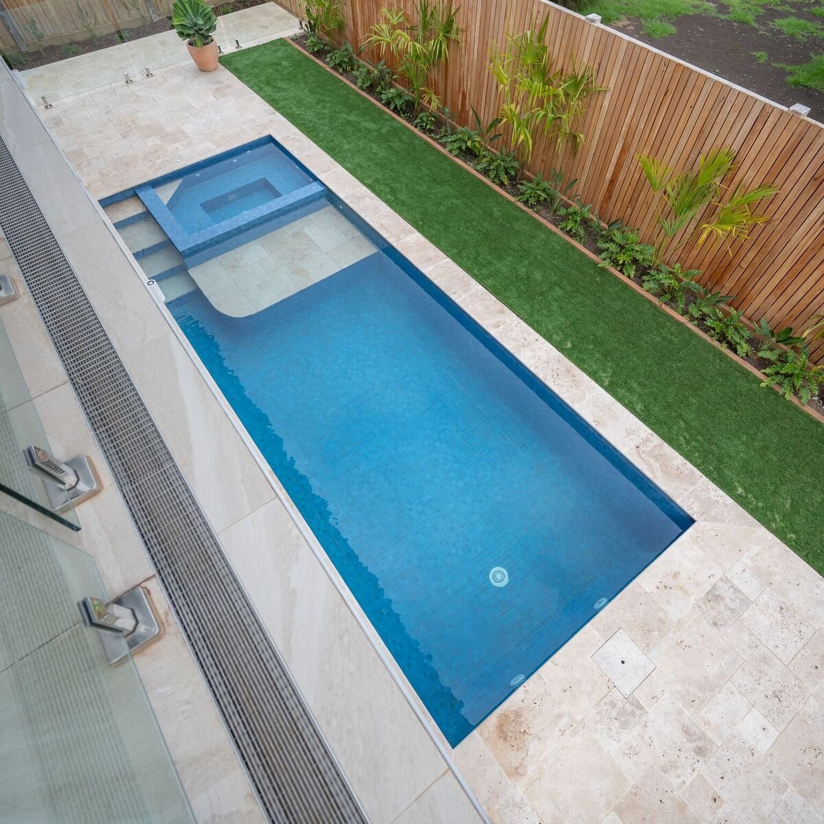 A modern backyard with a small rectangular swimming pool and a separate hot tub, surrounded by beige stone tiles, a narrow strip of artificial grass, and a wooden privacy fence lined with tropical plants.