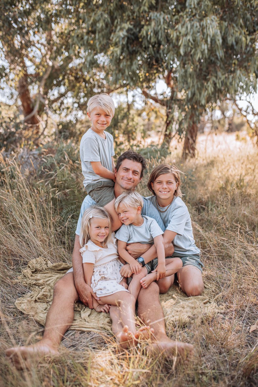 A man and five children sitting on a blanket outdoors in a grassy area with trees behind them, smiling and enjoying nature.