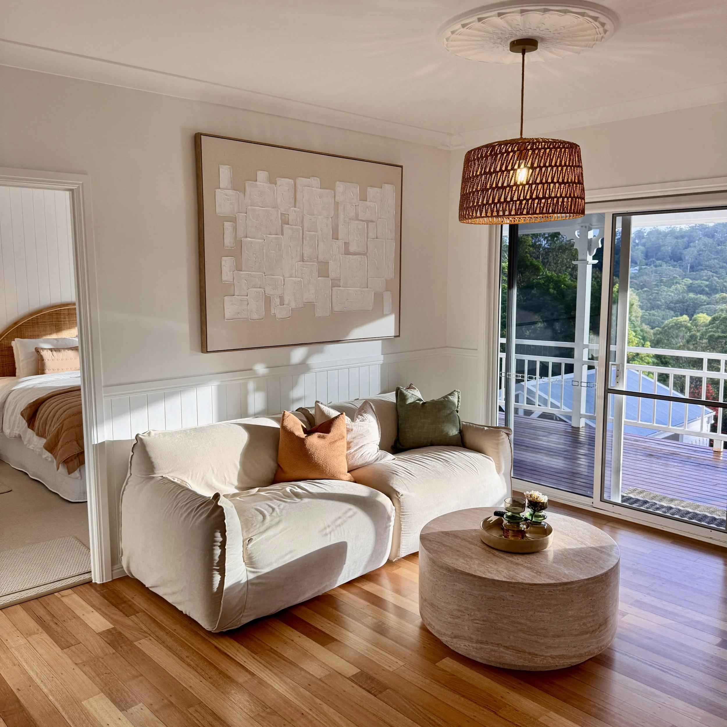 Living room with a beige sofa, colorful pillows, wooden coffee table, artwork on white wall, sliding glass door revealing a balcony with a view of a forested area.