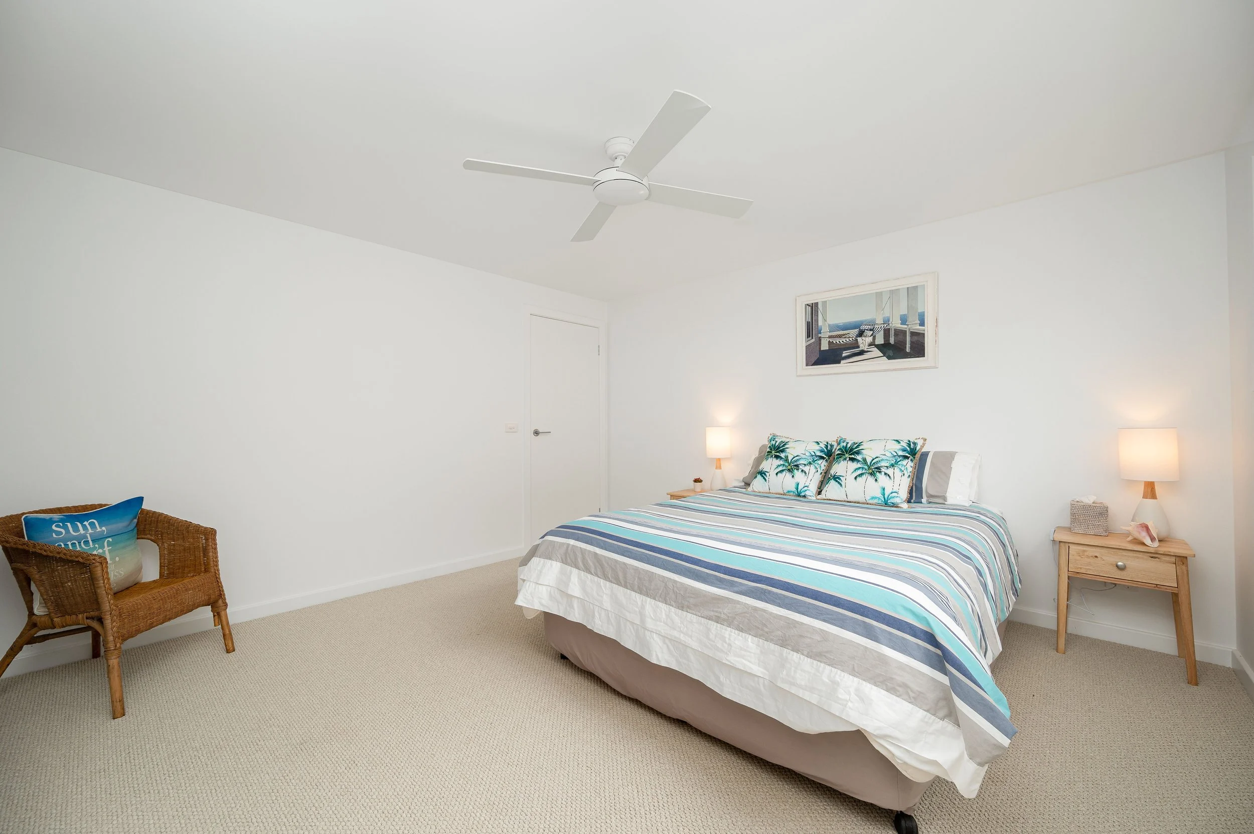 A bedroom with a bed featuring striped bedding and palm tree pillows, two bedside lamps, a wicker chair with a seashell pillow, and wall art of a beach scene, in a bright, minimalist style.